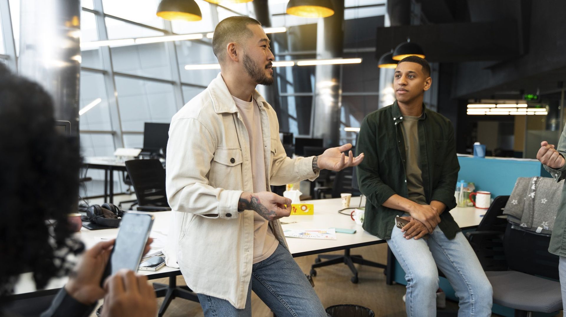 Two people talking in an office, one seated in a beige jacket and one standing in a dark green shirt.