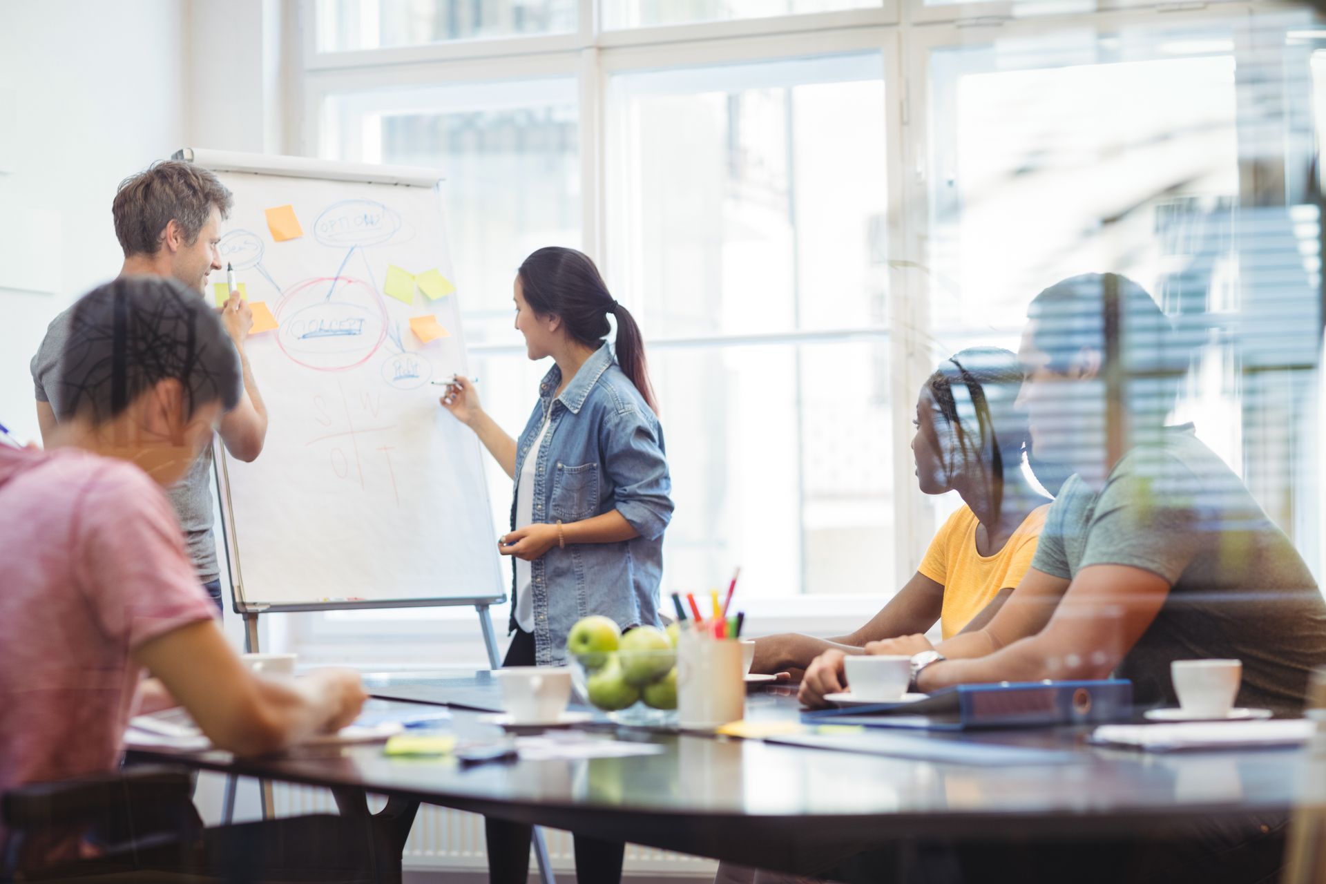 A group of people collaborating in a bright, modern office as one person points to a whiteboard with sticky notes.