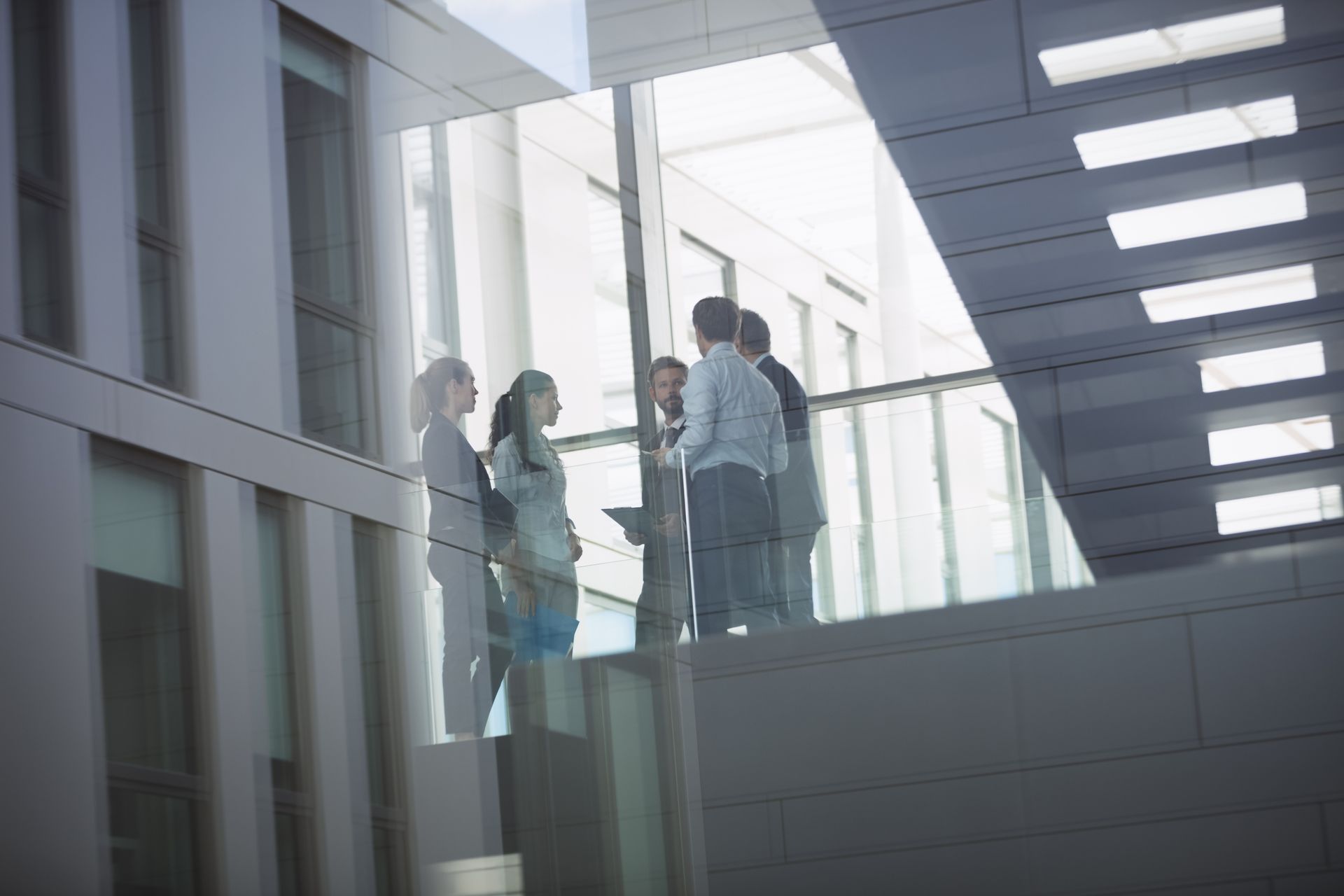 A group of professionals in business attire stand on a modern office bridge, engaged in conversation.