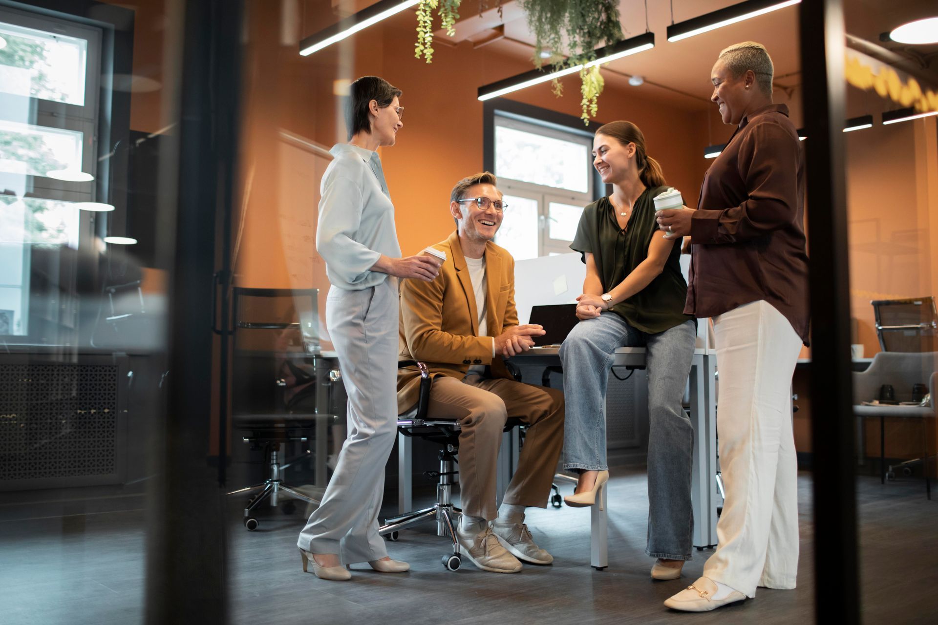 Four colleagues in an office setting stand and sit around a desk, talking and holding coffee cups.