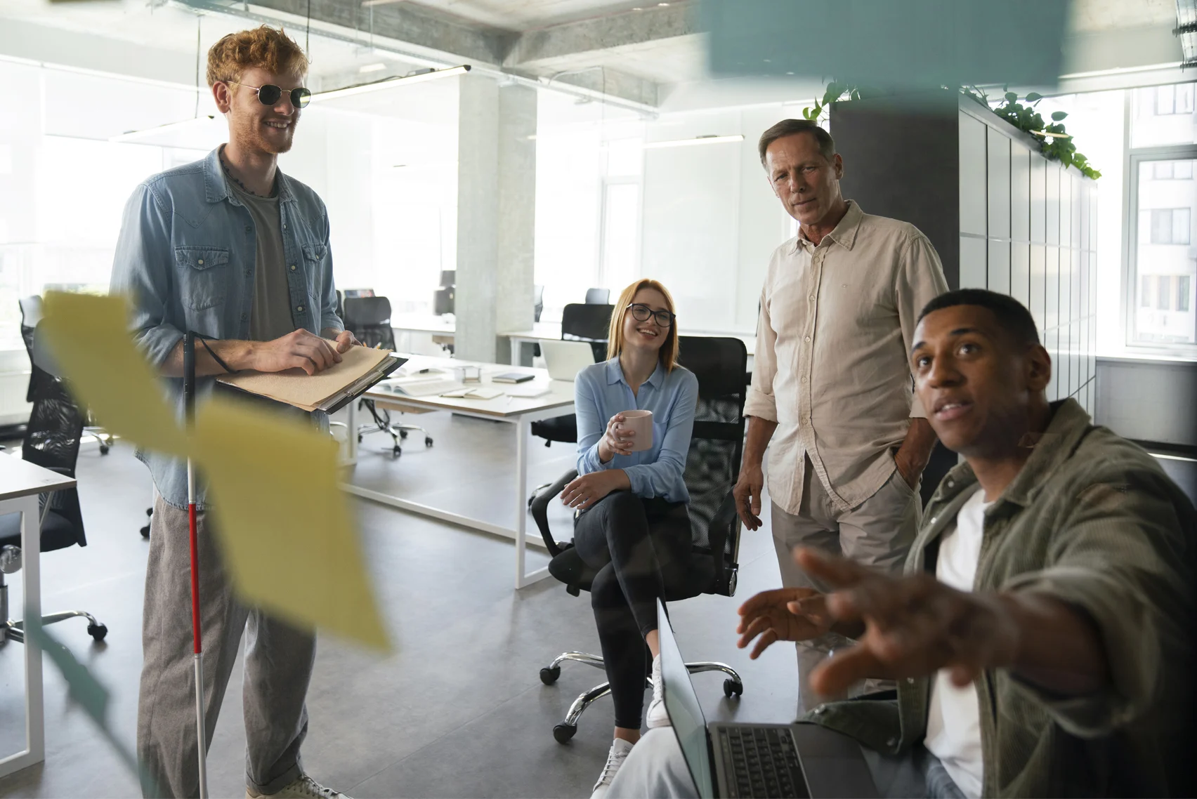 A group of colleagues collaborate in a modern office, pointing at notes and discussing a project while using a laptop.