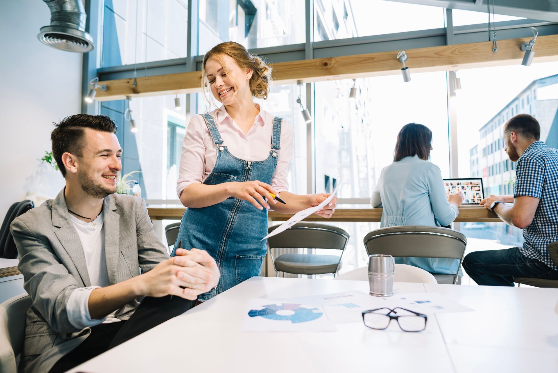 A woman in denim overalls discusses documents with a man in a blazer while others work at a sunlit office workspace.