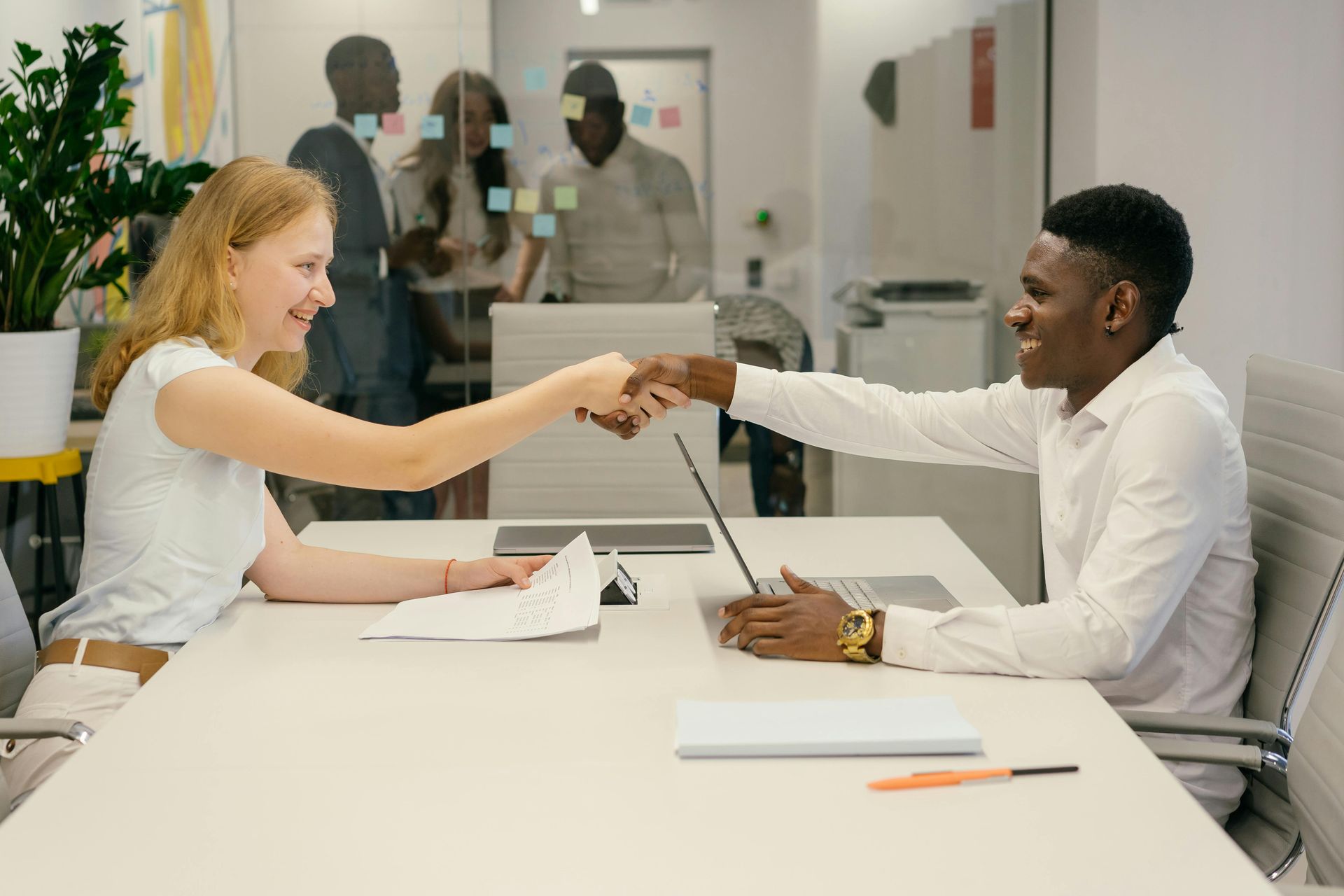 Two people shaking hands across a conference table in a bright office meeting room.