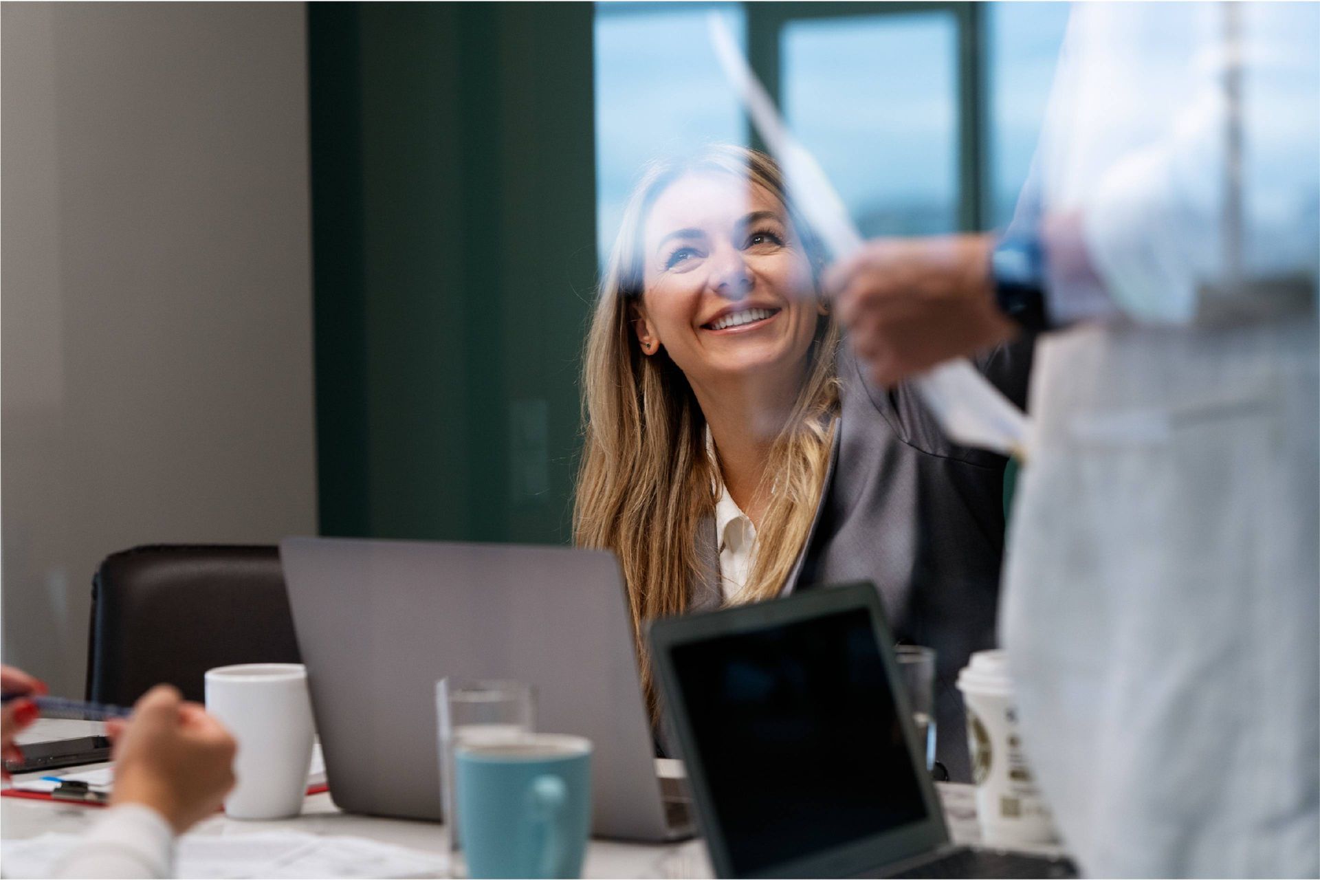 People in a meeting room with a smiling woman behind a glass partition and laptops on the table