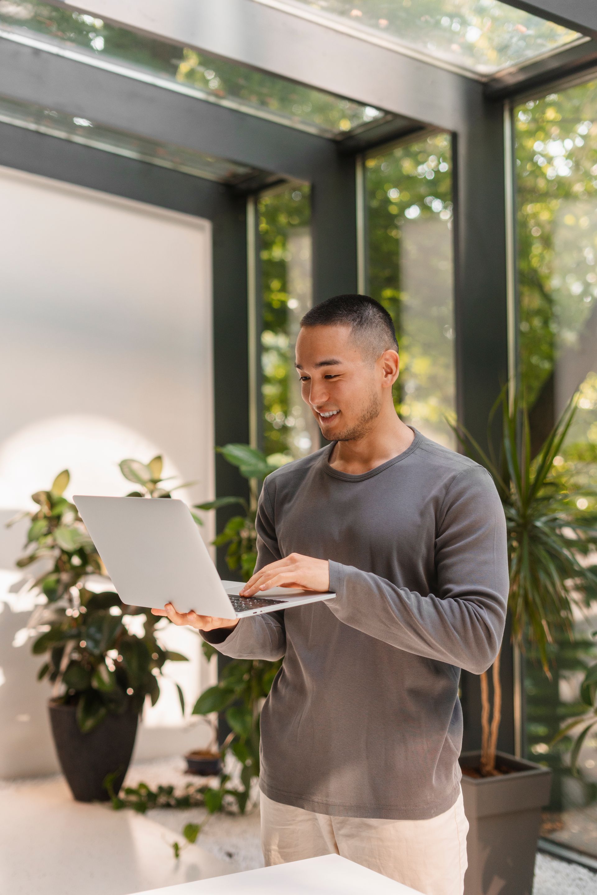 A person in a long-sleeved gray shirt standing in a sunlit atrium and using a laptop.