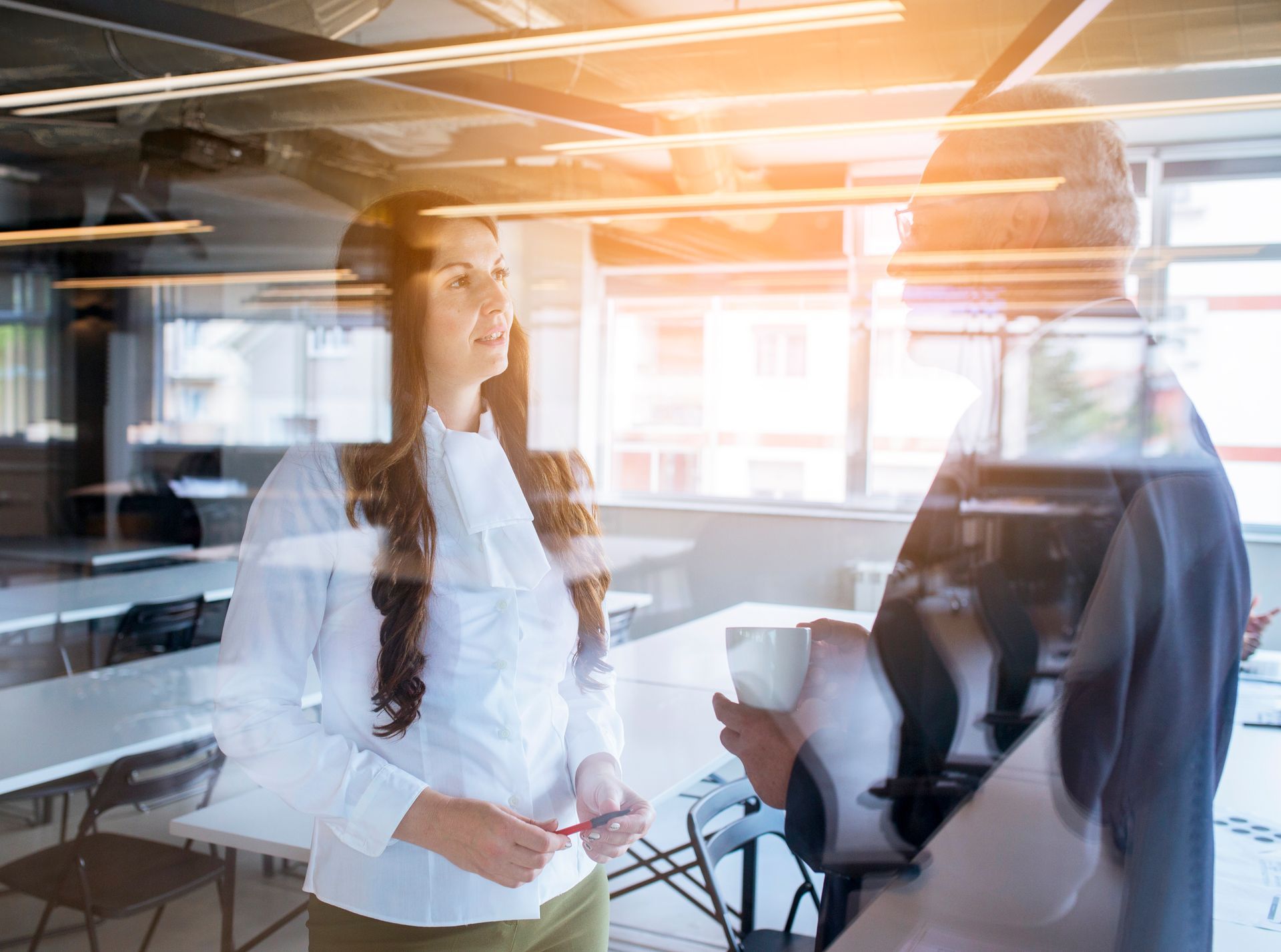 Two people in business attire stand in a sunlit office, facing each other and appearing to have a professional discussion.