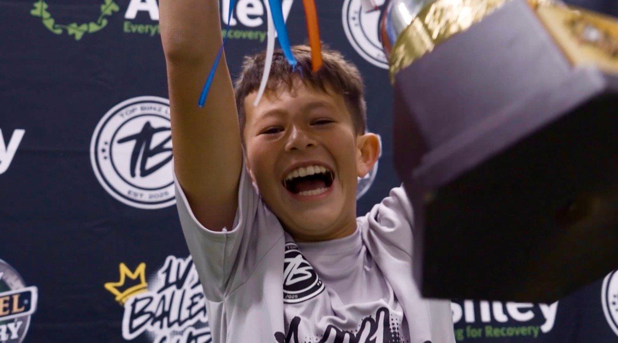 Boy with trophy raises arms, smiling. Indoor soccer event banner in background.