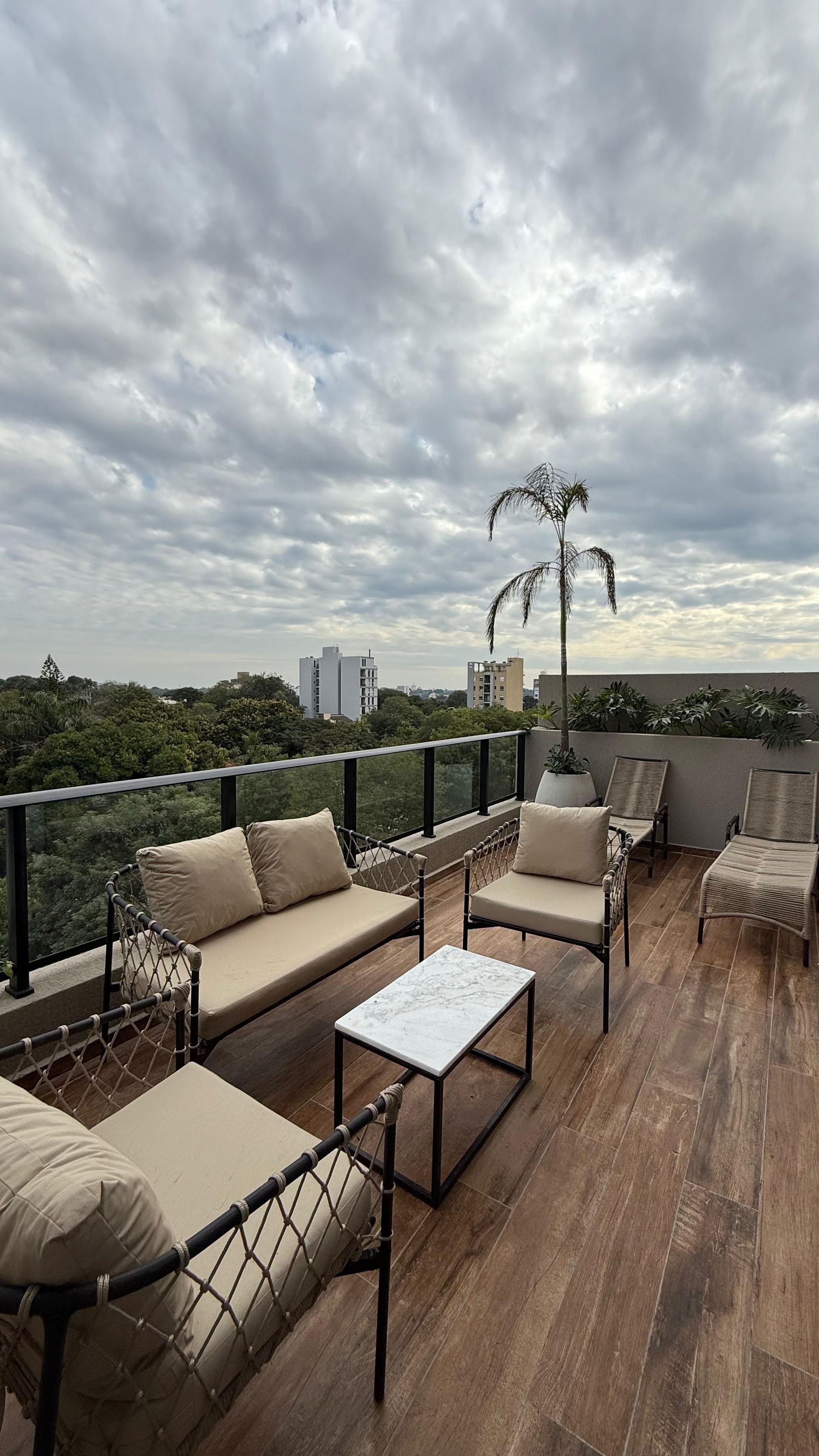 Patio en la azotea con muebles beige, estructura de metal, mesa de centro de mármol y cielo nublado.