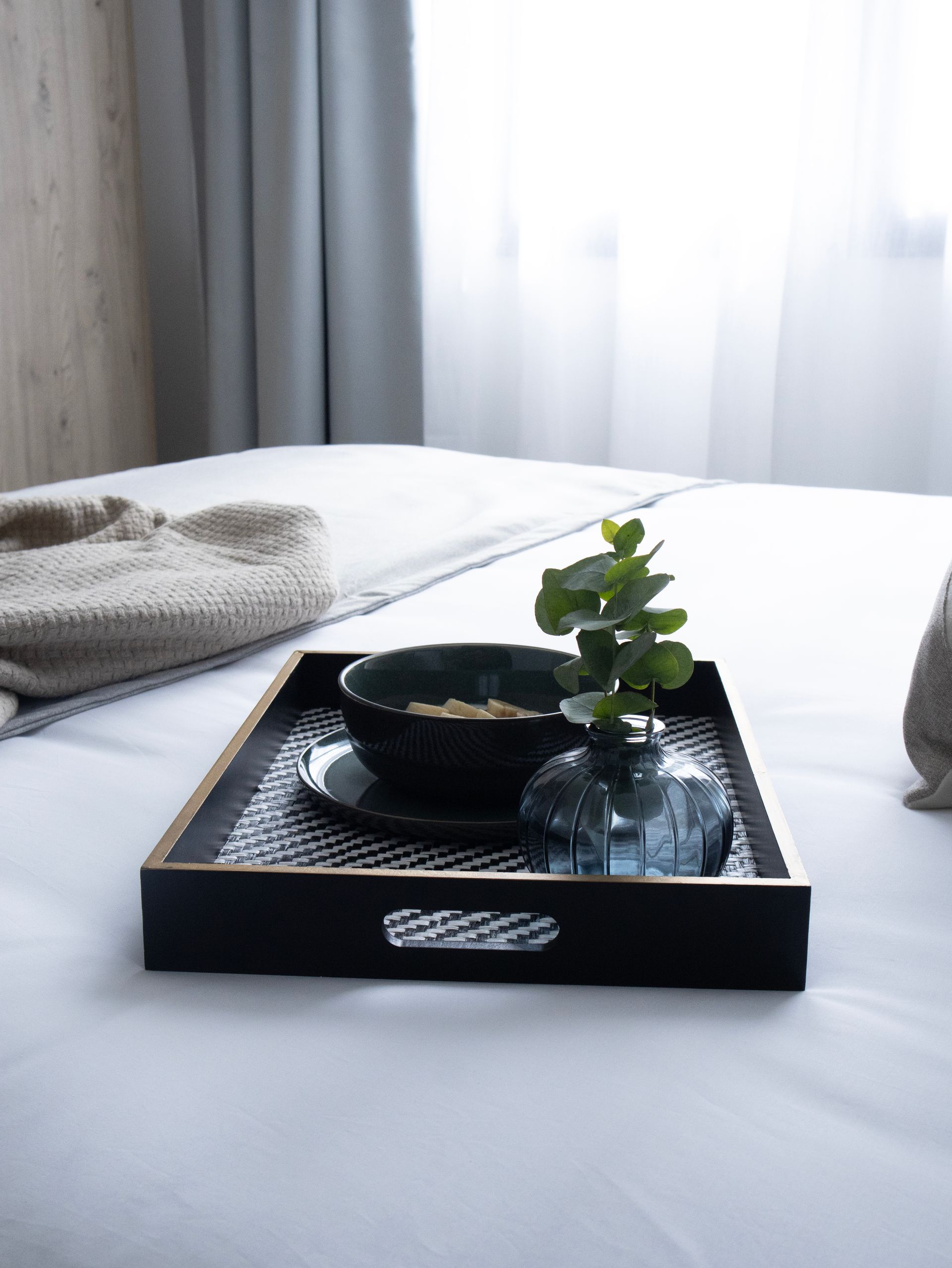 Bandeja de desayuno en la cama: bol de cereales, pequeño jarrón con plantas verdes, tonos neutros.
