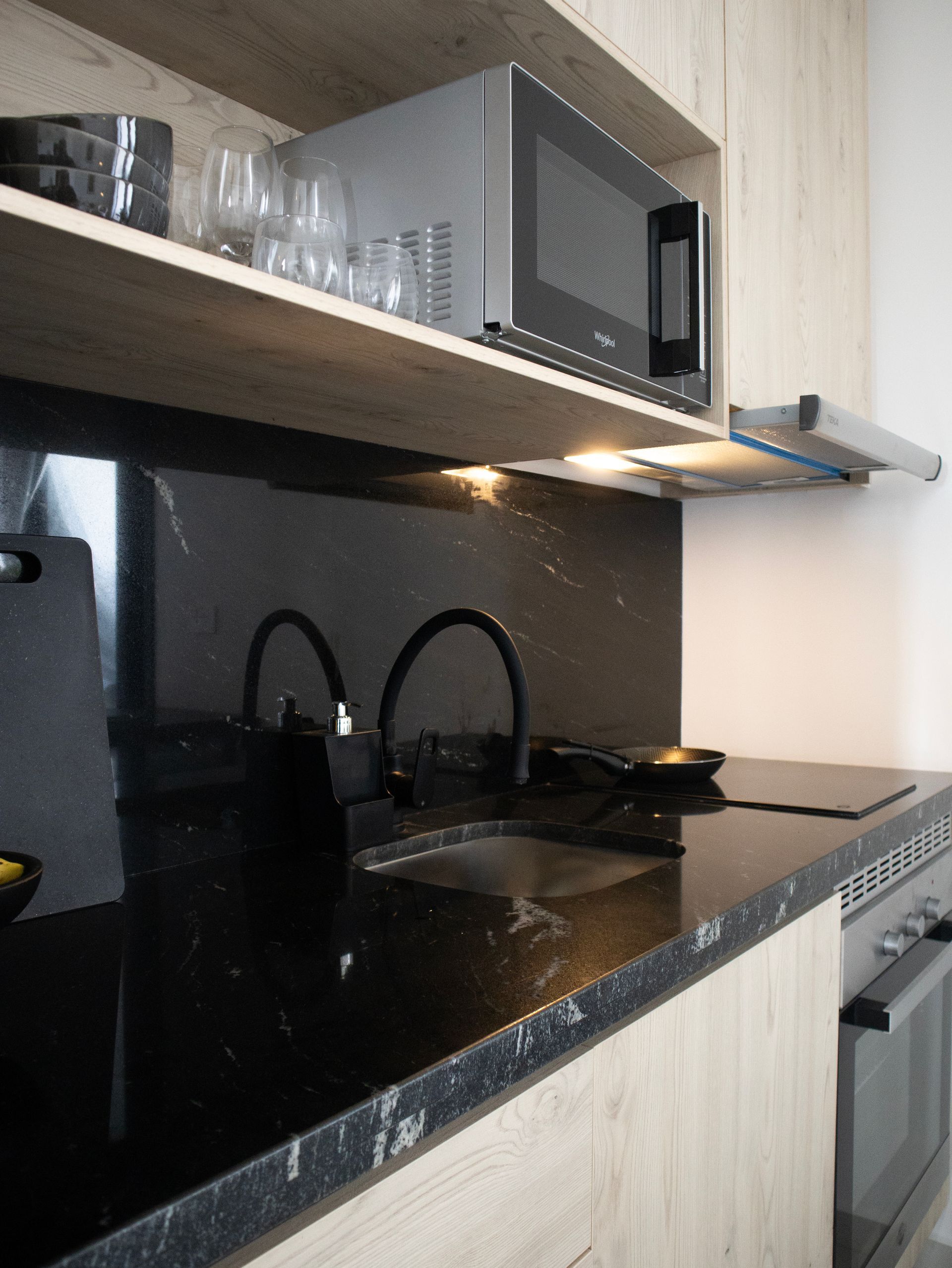 Encimera negra con fregadero y anafe, microondas sobre estante, grifo negro e iluminación de cocina.