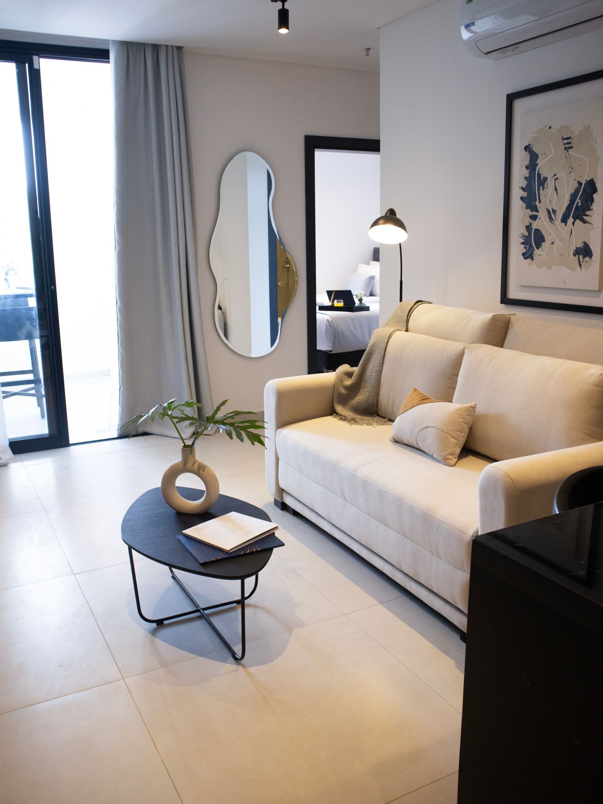 Sala de estar con sofá blanco, mesa de café negra y espejo grande; una puerta conduce a un dormitorio.