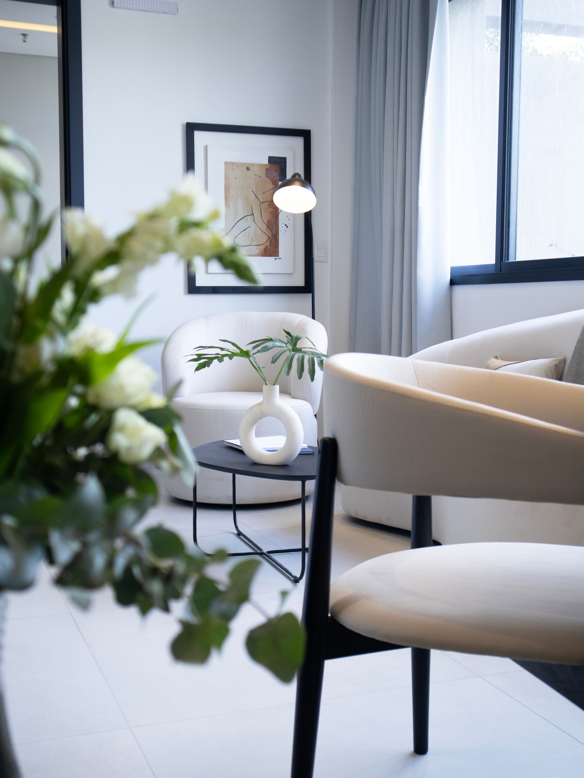 Acogedor espacio de estar con sillones blancos, una mesa pequeña y un cuadro, cerca de una ventana y flores.