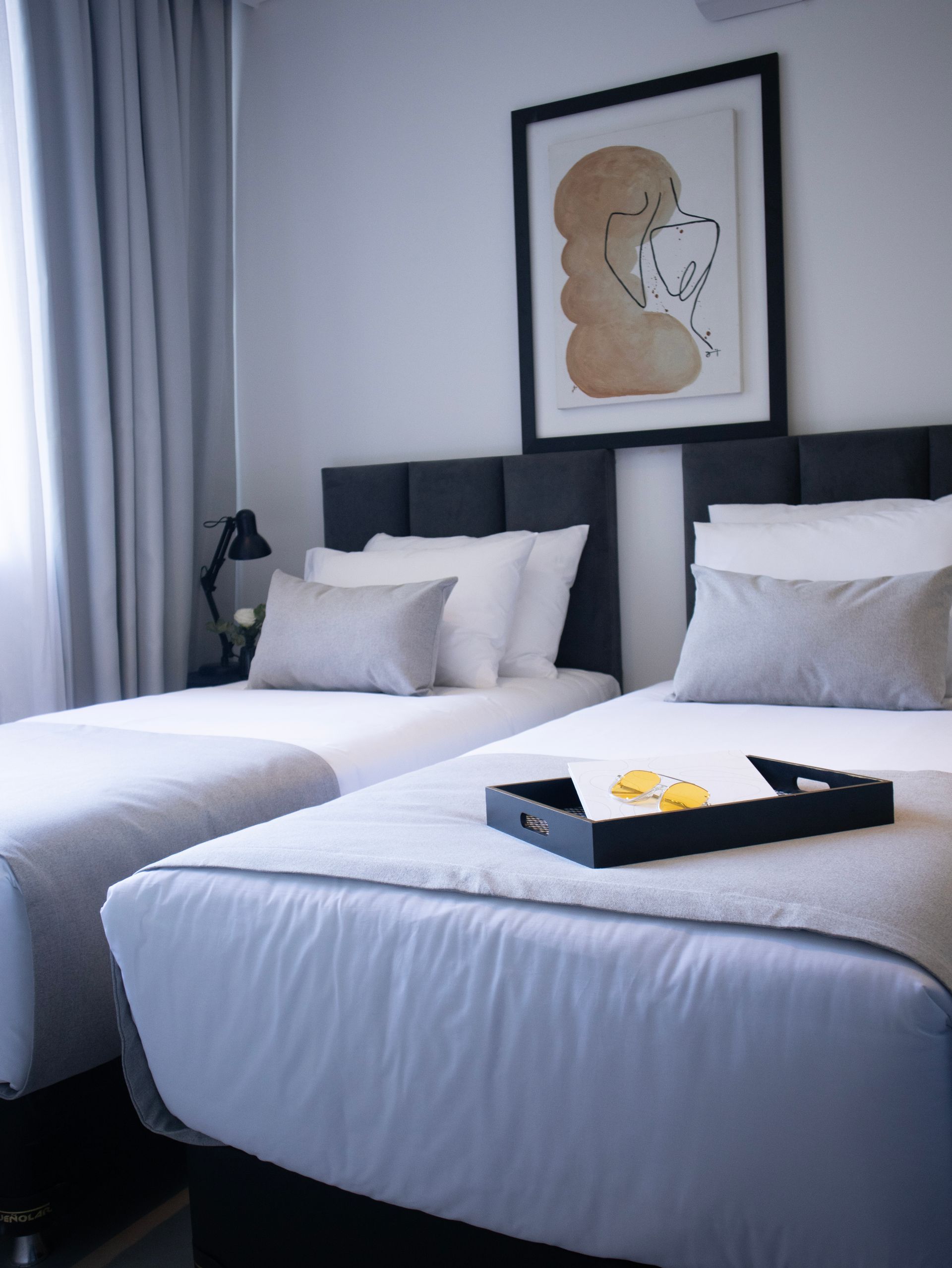 Dos camas individuales con sábanas blancas y almohadas grises en un dormitorio con una obra de arte enmarcada.