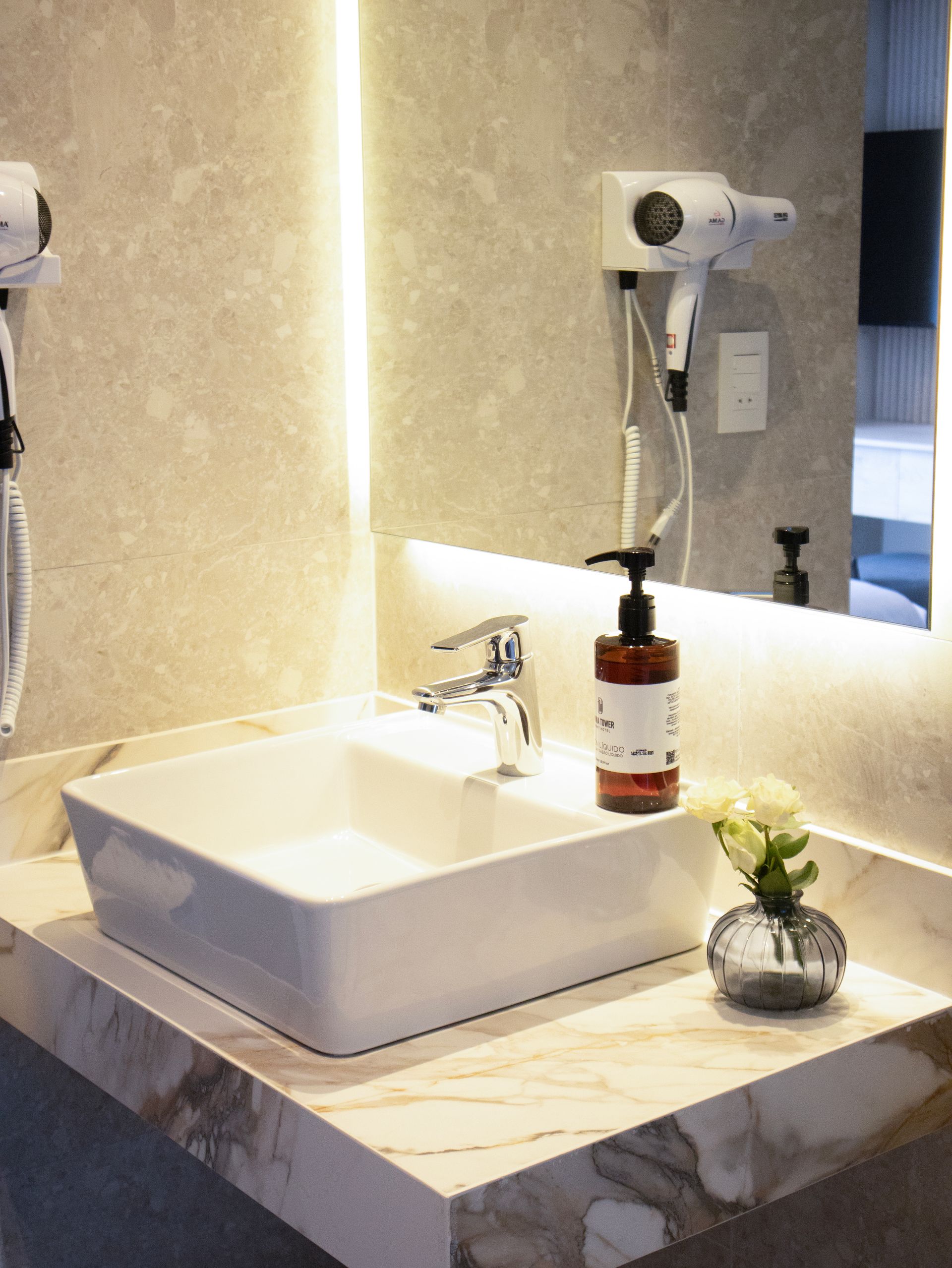 Lavabo de baño con jabón, flores y secador de pelo sobre encimera de mármol.