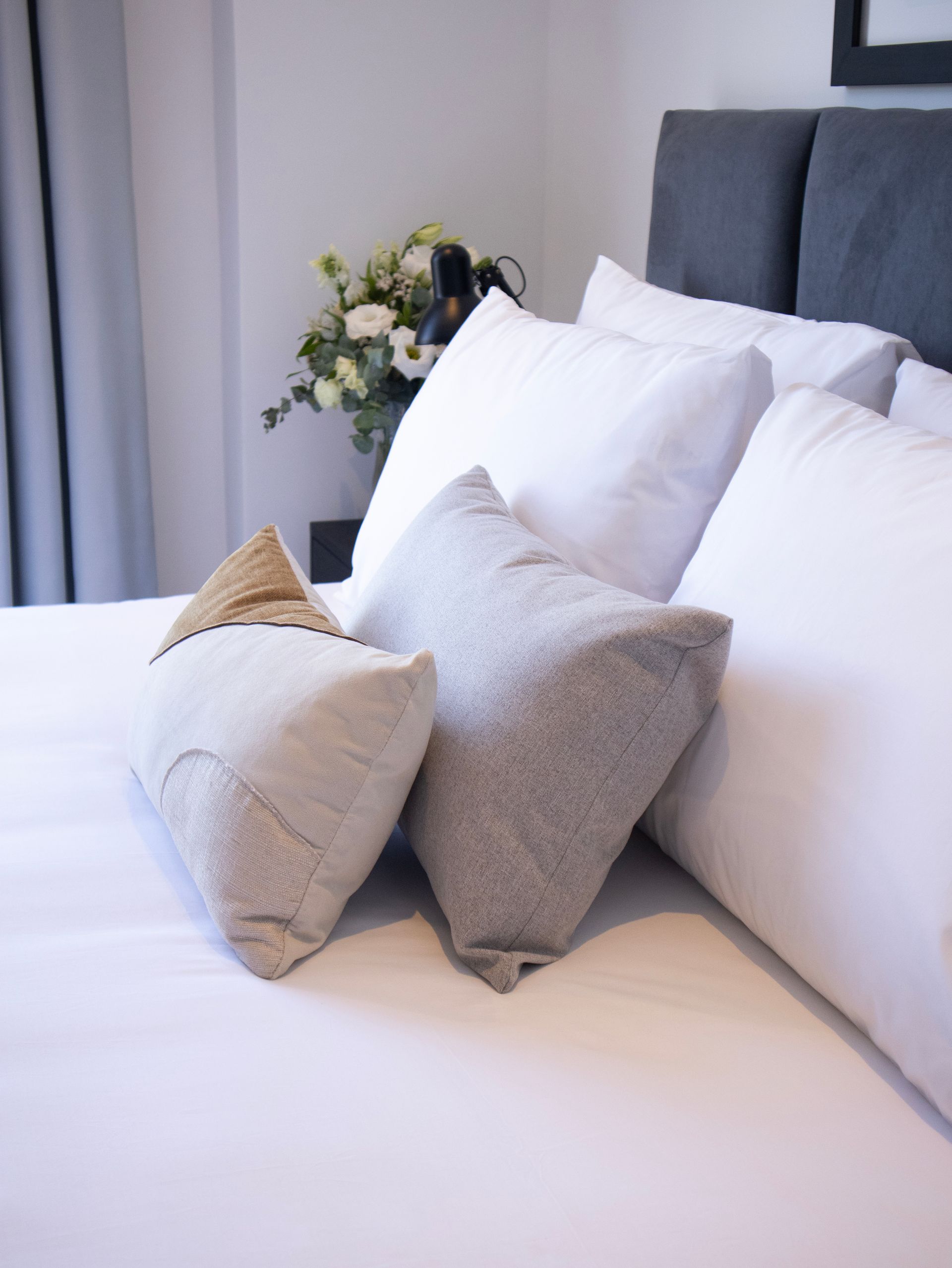 Cama con ropa de cama blanca y almohadas decorativas, junto a un ramo de flores.