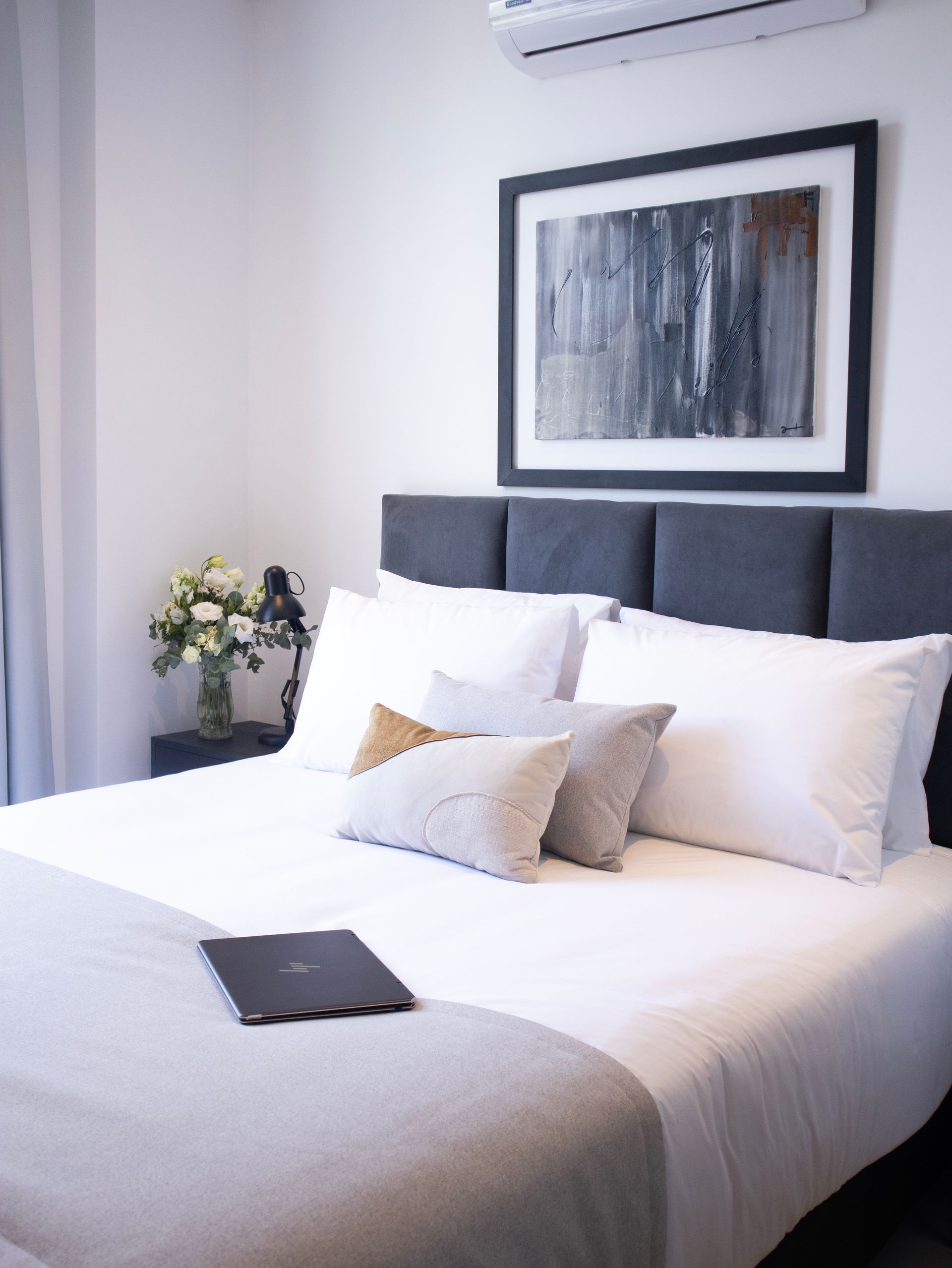 Dormitorio con cama, almohadas, obras de arte y flores. Portátil sobre la cama.