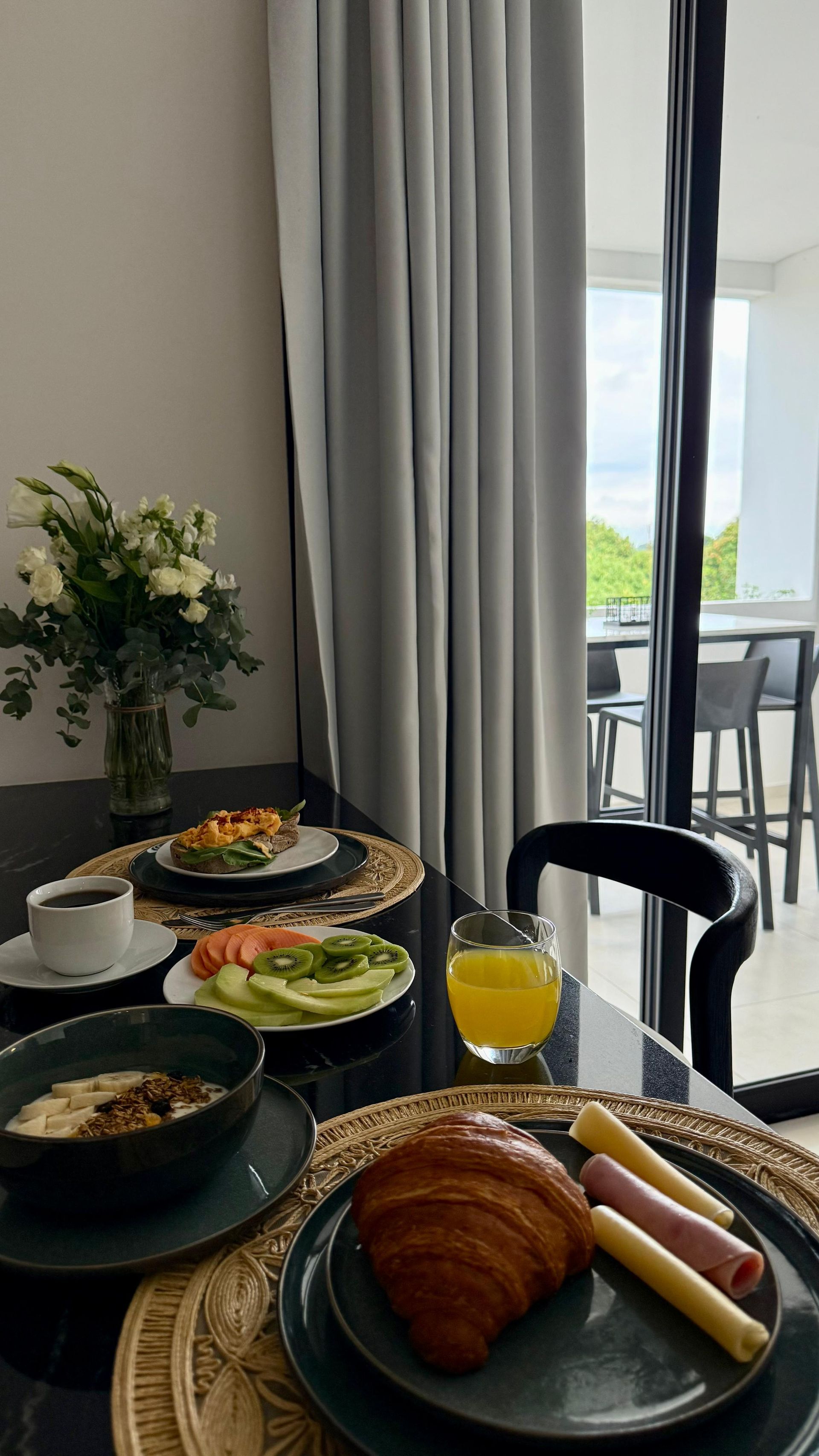 Mesa de desayuno: croissant, jamón y queso, avena, tostadas, fruta, jugo, flores.