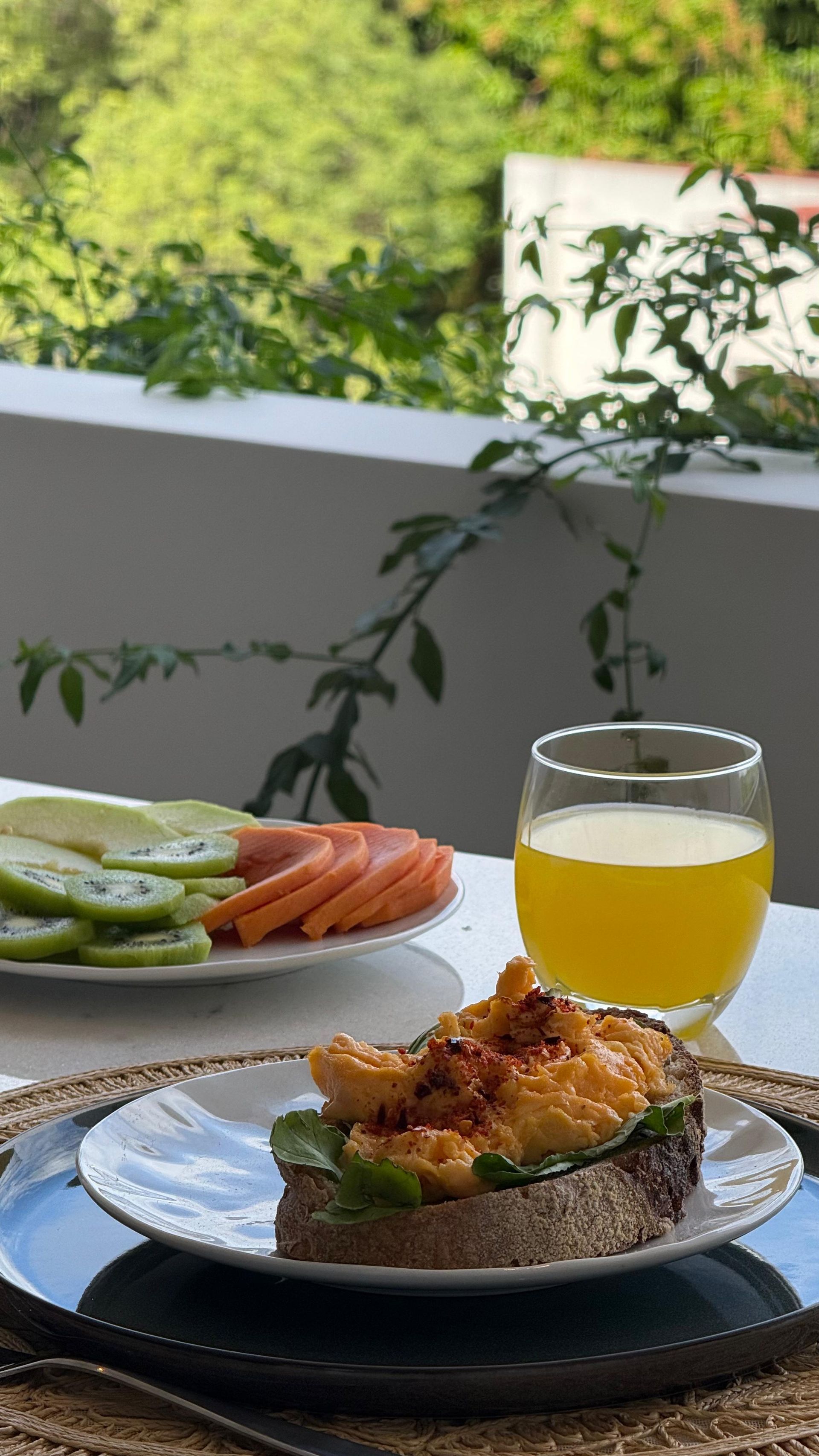 Desayuno en un balcón: tostadas con huevos revueltos, plato de frutas, vaso de jugo de naranja, vegetación de fondo.