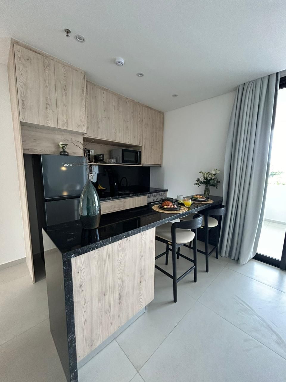 Cocina moderna con gabinetes de madera clara, encimera negra y comedor con dos taburetes.