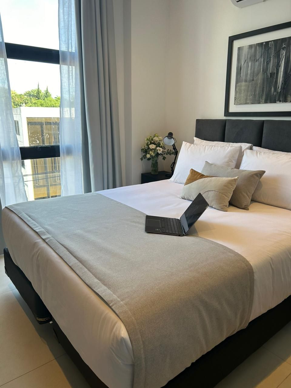 Dormitorio con cama, portátil, ventana, obras de arte y flores. Tonos blancos, grises y oscuros.