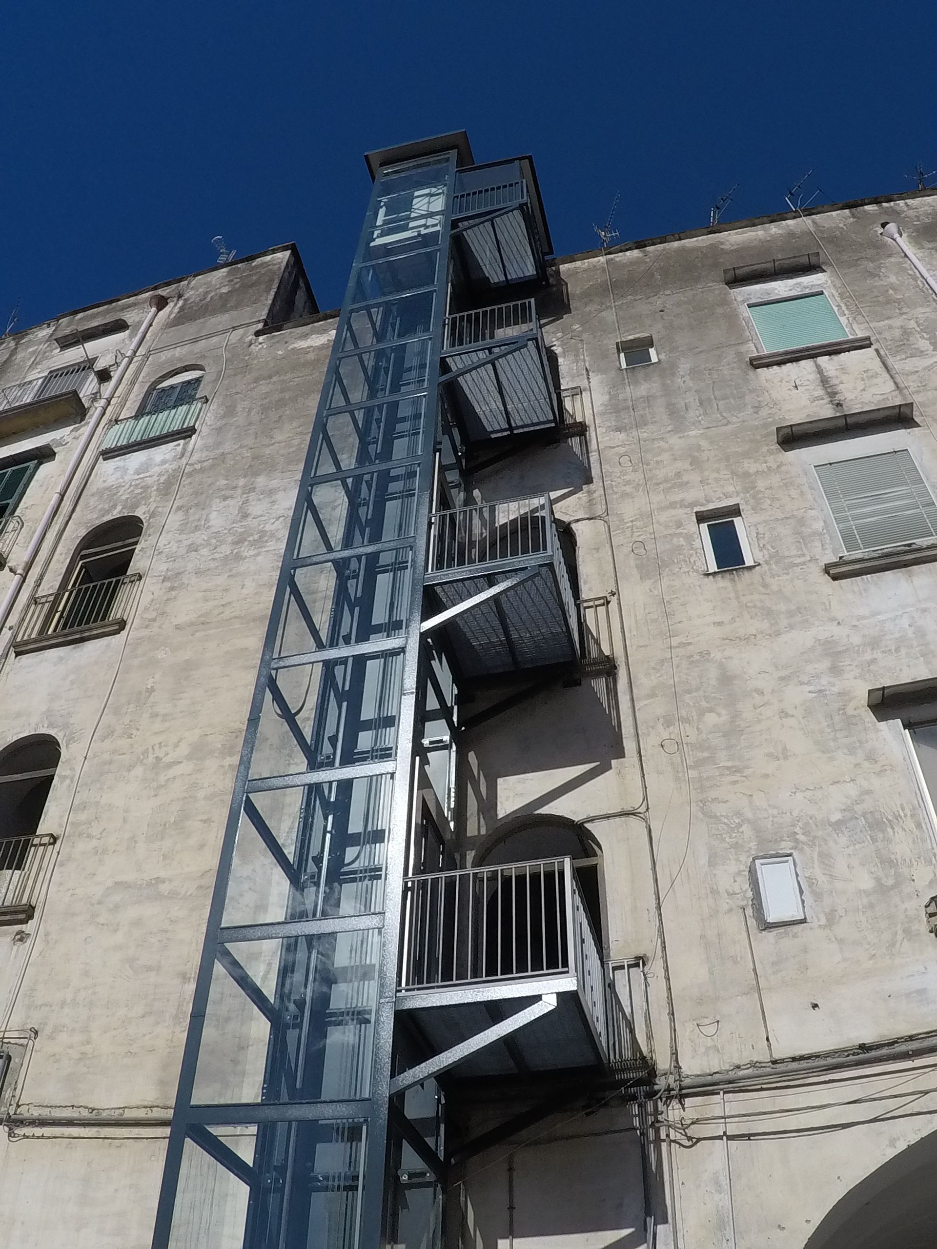 Ascensore esterno collegato a un edificio usurato con più piani e piattaforme metalliche. Cielo azzurro brillante.