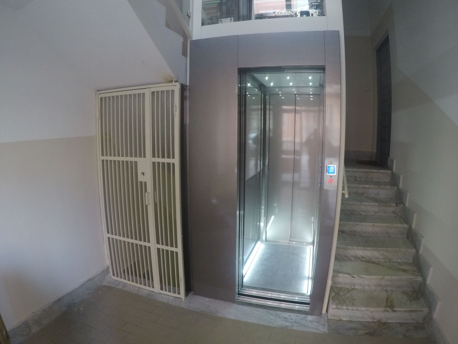 Ascensore in un edificio con cancello metallico aperto a sinistra e scale a destra. La porta dell'ascensore è aperta.