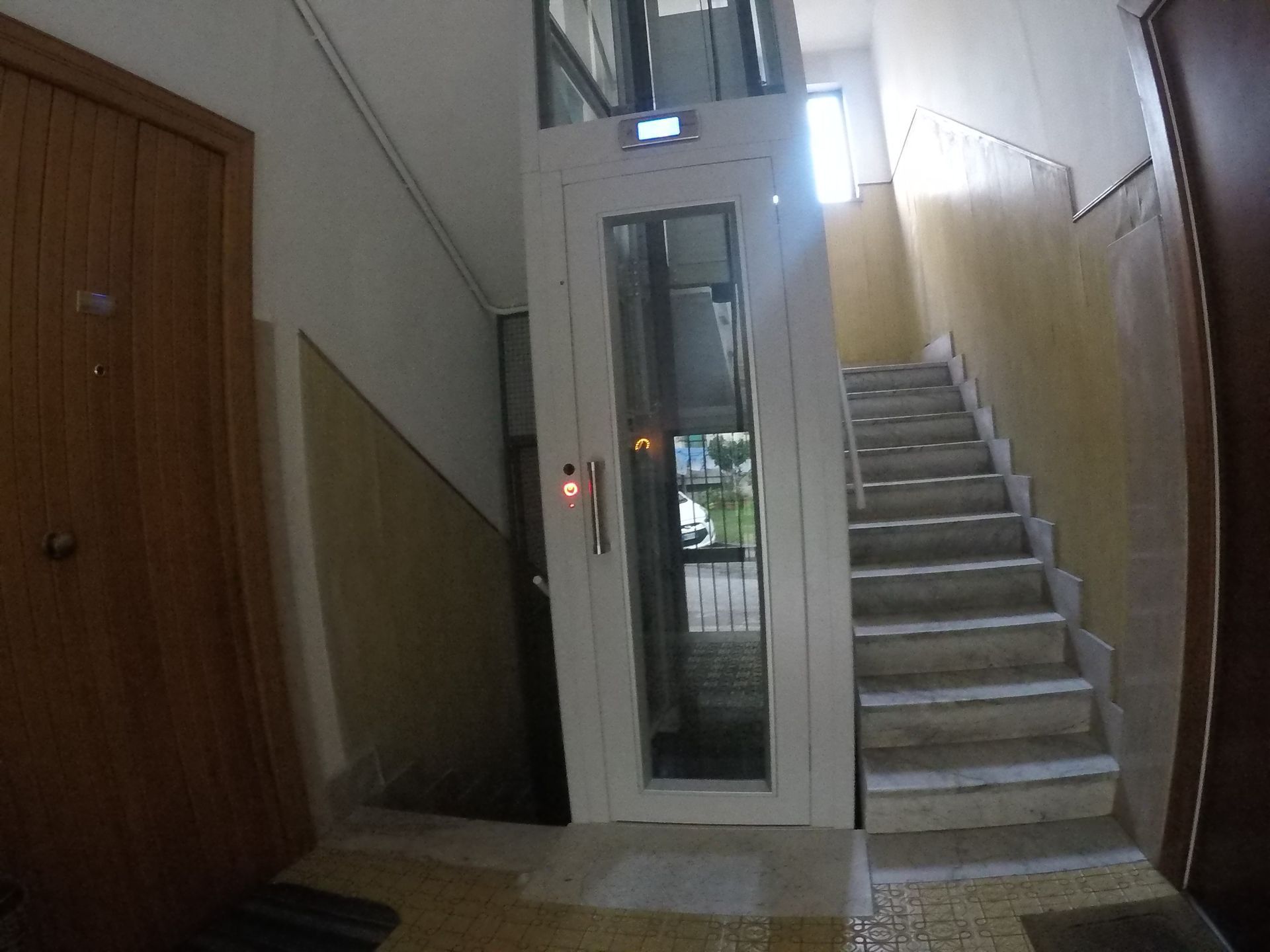 Ascensore installato accanto a una scala nel corridoio di un edificio. Ascensore bianco con pannelli in vetro e porta sulla destra.