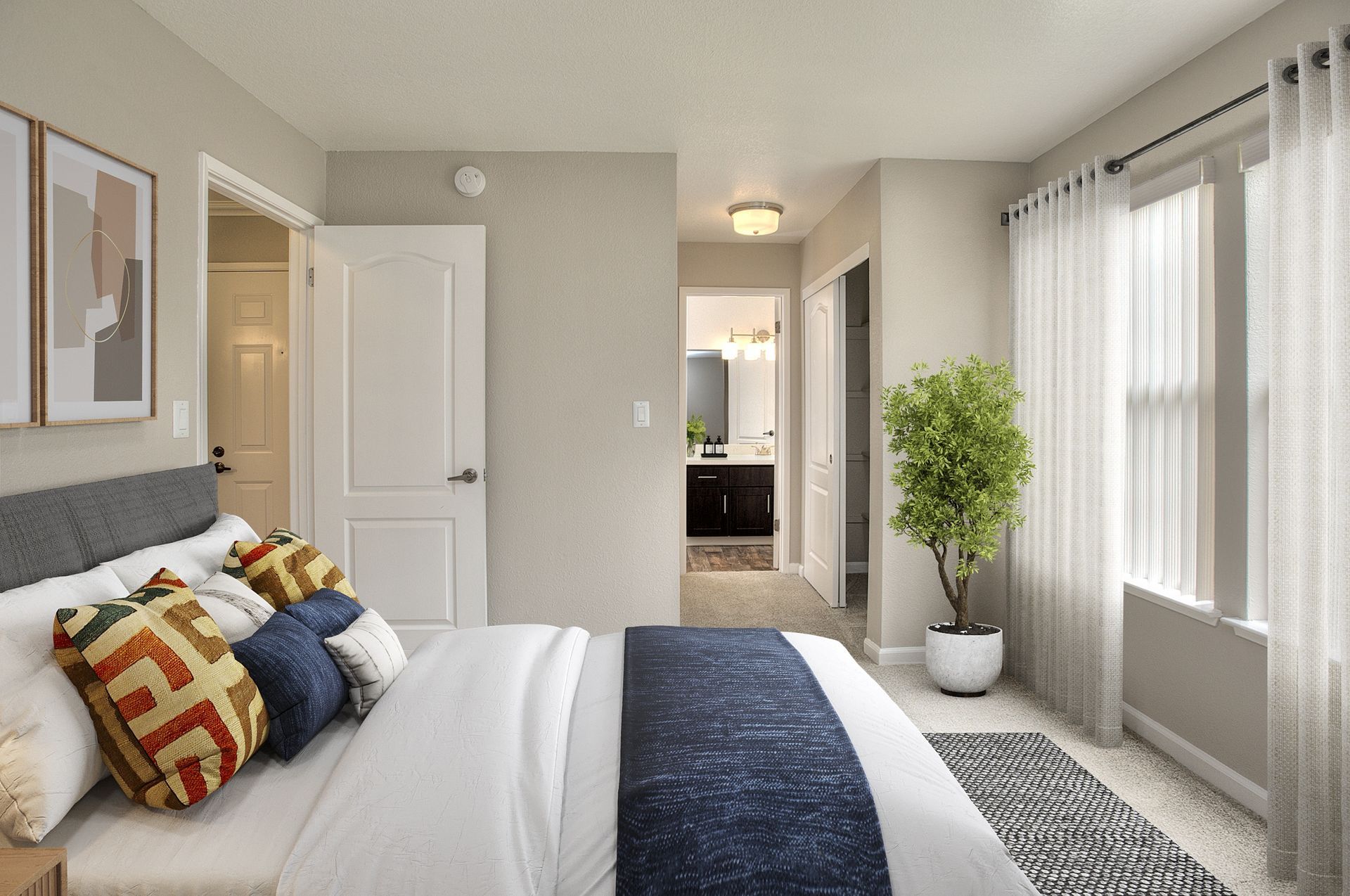 Bedroom with a made bed, decorative pillows, a large window with sheer curtains, and a doorway to a bathroom.