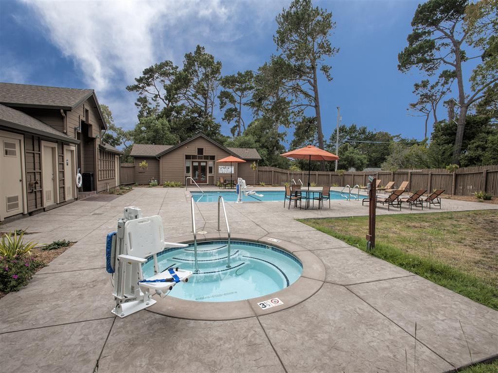 Outdoor apartment community pool area with a hot tub, lounge chairs, and umbrellas on a paved deck.