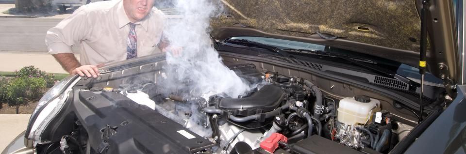 A person in business attire looks down with concern at a smoking vehicle engine with the hood open.