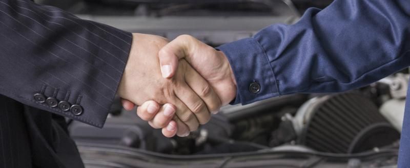A business person in a suit and a worker in blue uniform shaking hands in front of an open car engine.