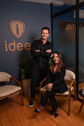 Duas pessoas sorrindo em um escritório com um logotipo na parede. O homem está em pé com os braços cruzados, a mulher está sentada.