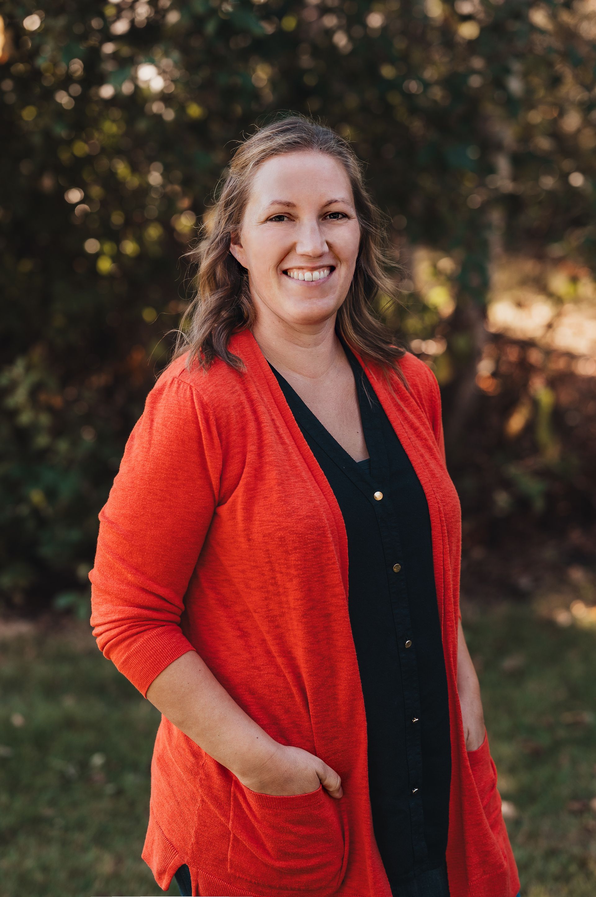 Woman in red cardigan smiles outdoors, hands in pockets.