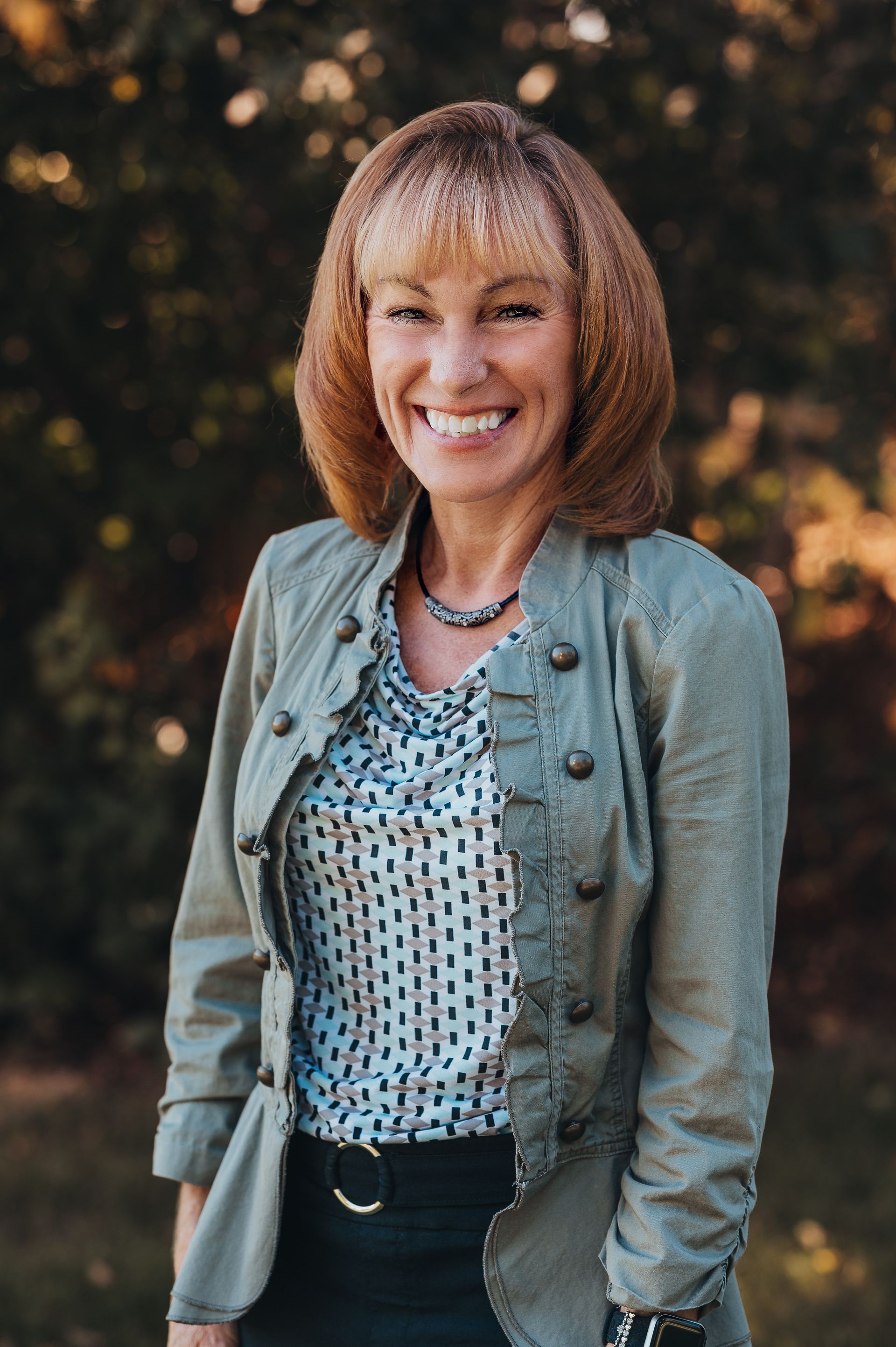Woman smiling, wearing a green jacket over a printed blouse, standing outside.
