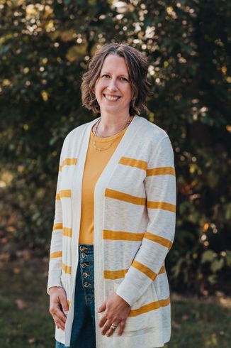 Woman with curly hair smiles, wearing a yellow shirt and striped cardigan, standing outdoors.