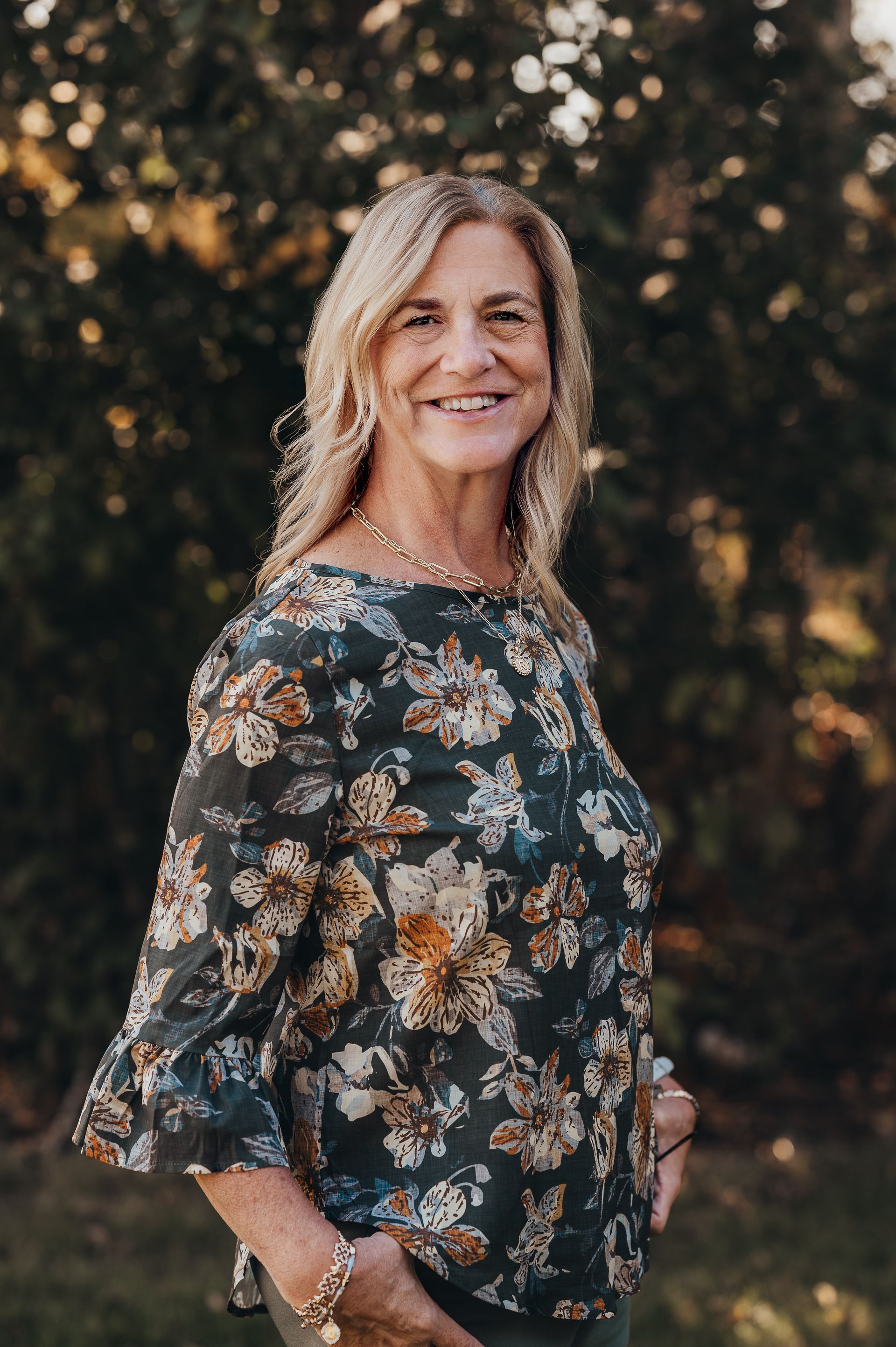 Woman in floral blouse smiles outdoors, hands in pockets.