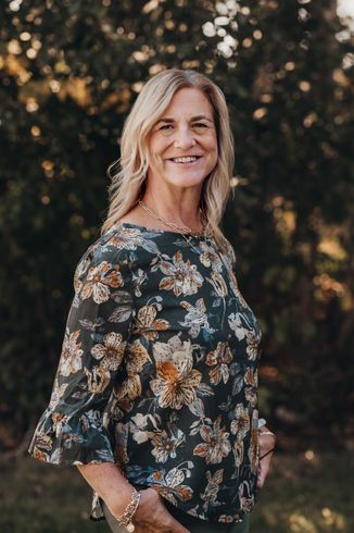 Woman in floral blouse smiles outdoors, hands in pockets.