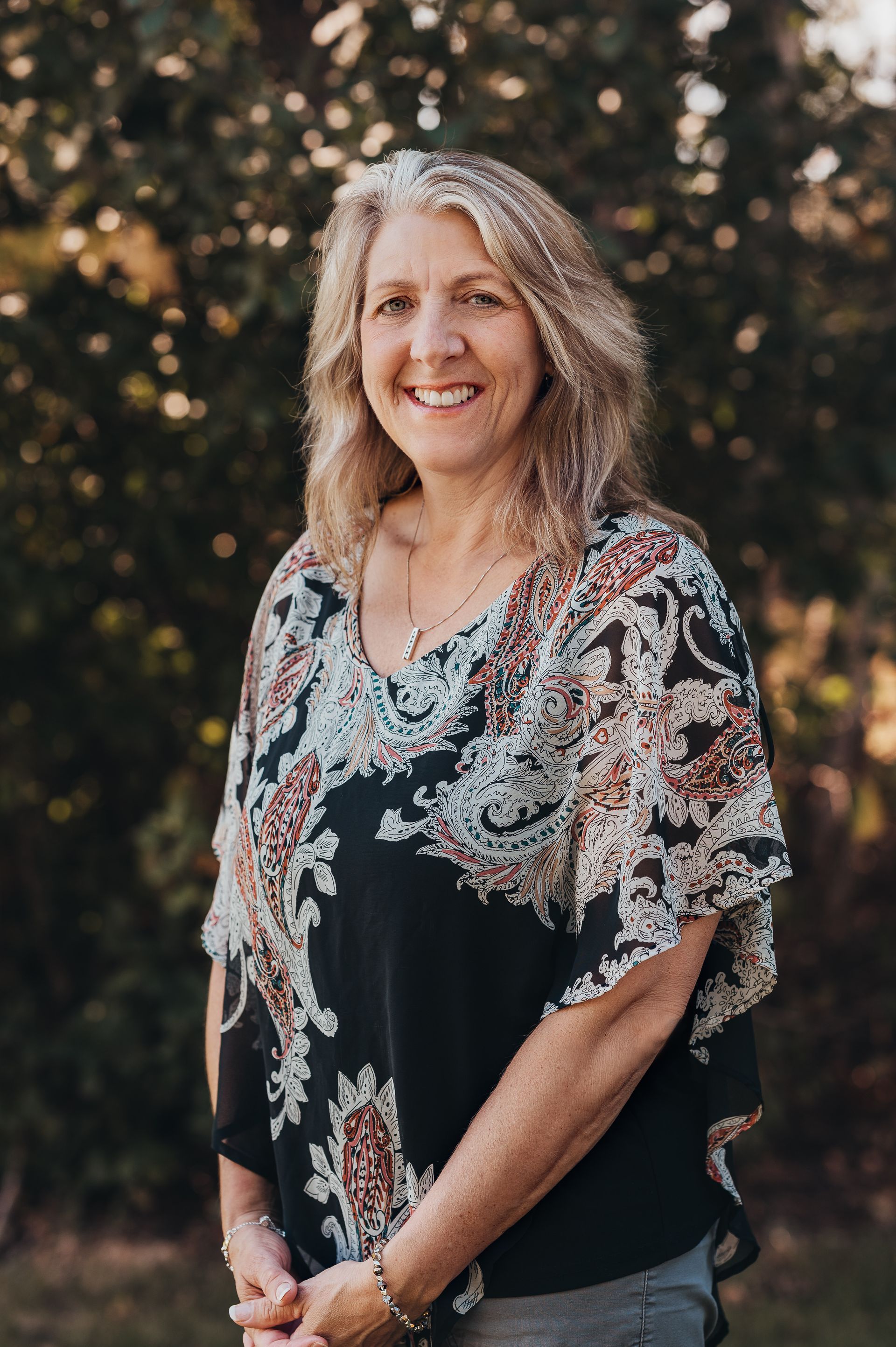 Woman with blonde hair wearing a patterned top, smiling outdoors.