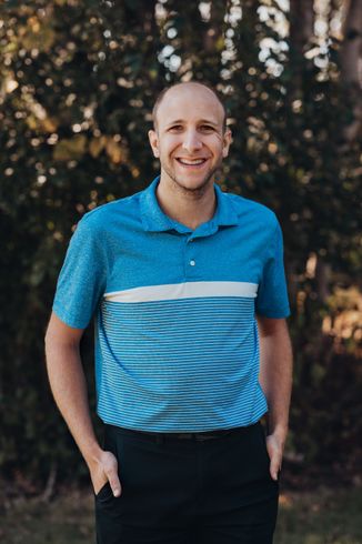 Man in blue patterned shirt smiles, hands in pockets; outdoor setting.