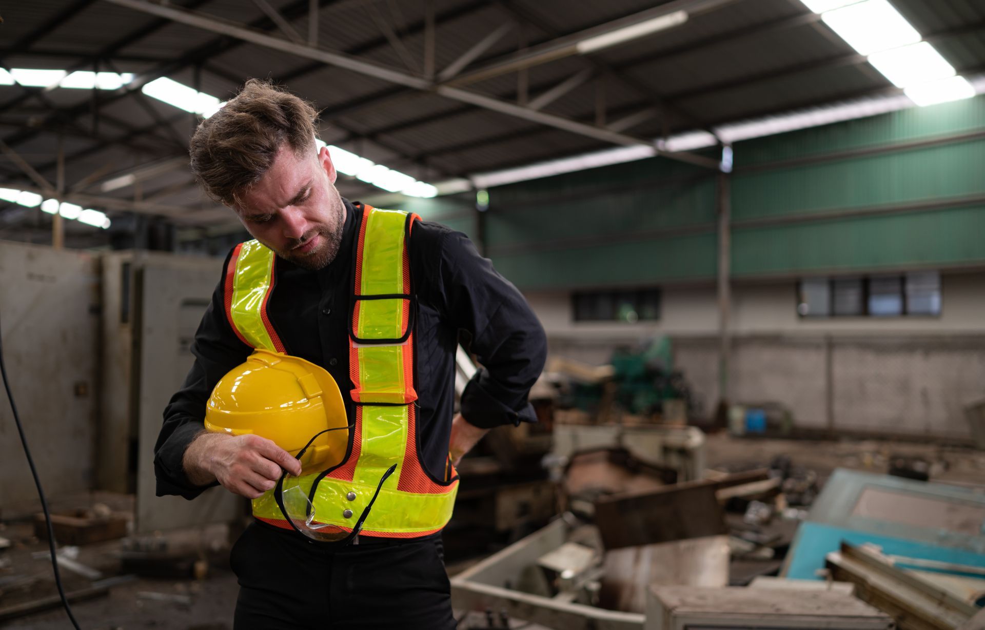 Man in safety vest holding yellow hard hat, inspecting his lower back in a cluttered industrial setting.