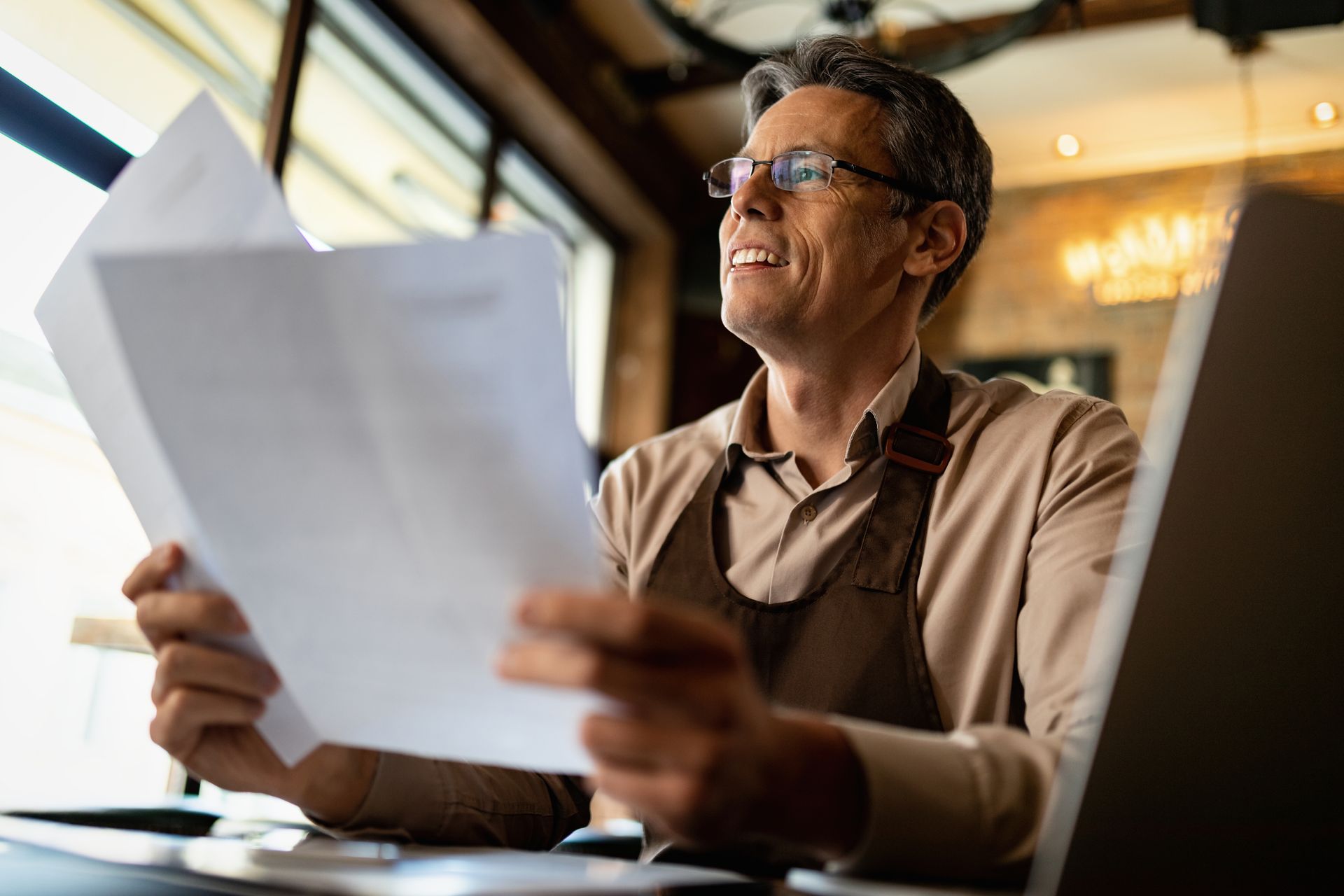 Man wearing glasses and apron reviews papers near a laptop, smiling in a cafe.