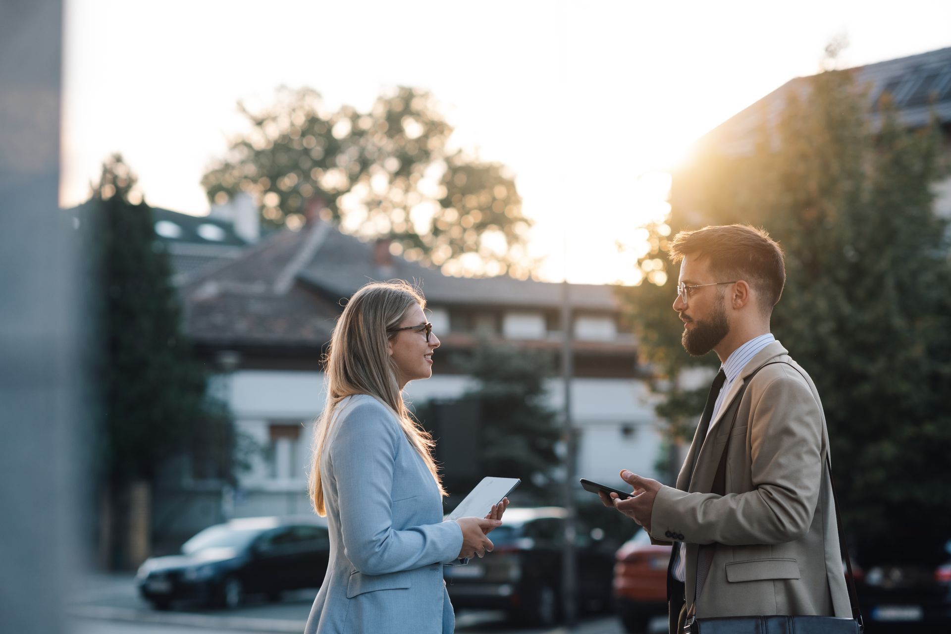 Two people in business attire conversing outdoors; sunlight in background; one holding a tablet, the other a phone.
