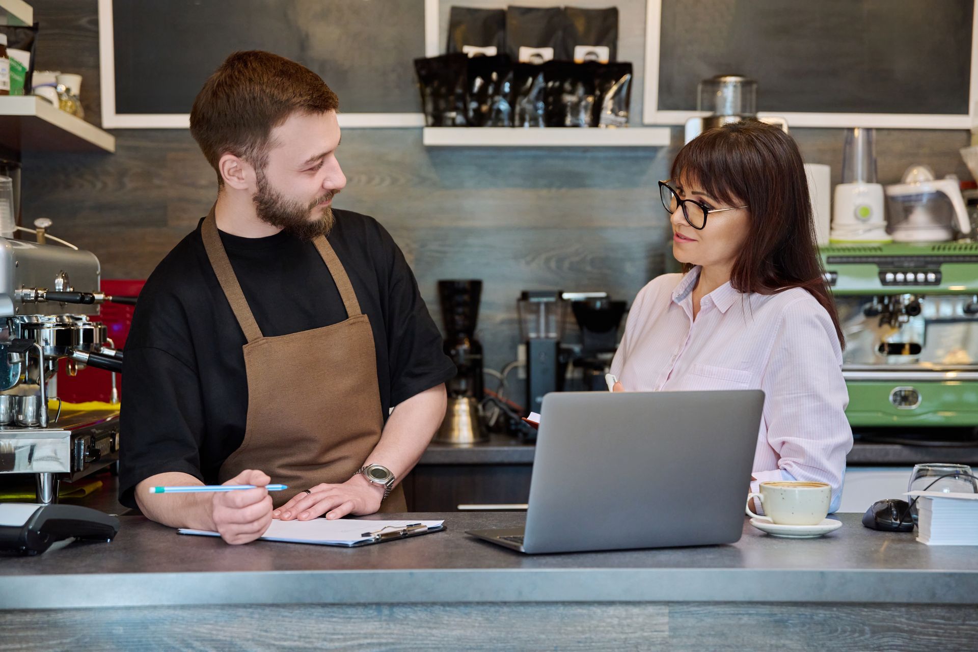 A barista and a woman with glasses, discussing while looking at a laptop and clipboard at a coffee shop counter.
