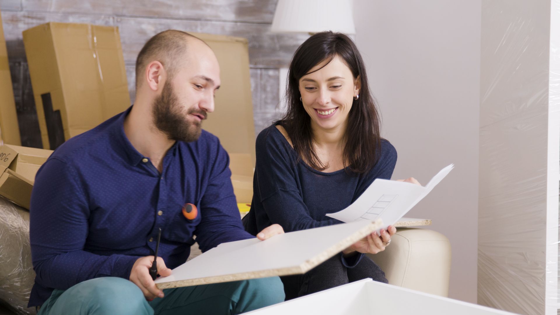 Couple assembling furniture, looking at instructions in a new home, surrounded by boxes and packing materials.