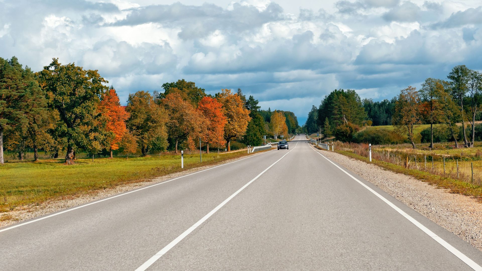 Paved road through a landscape with trees displaying autumn colors under a cloudy sky.