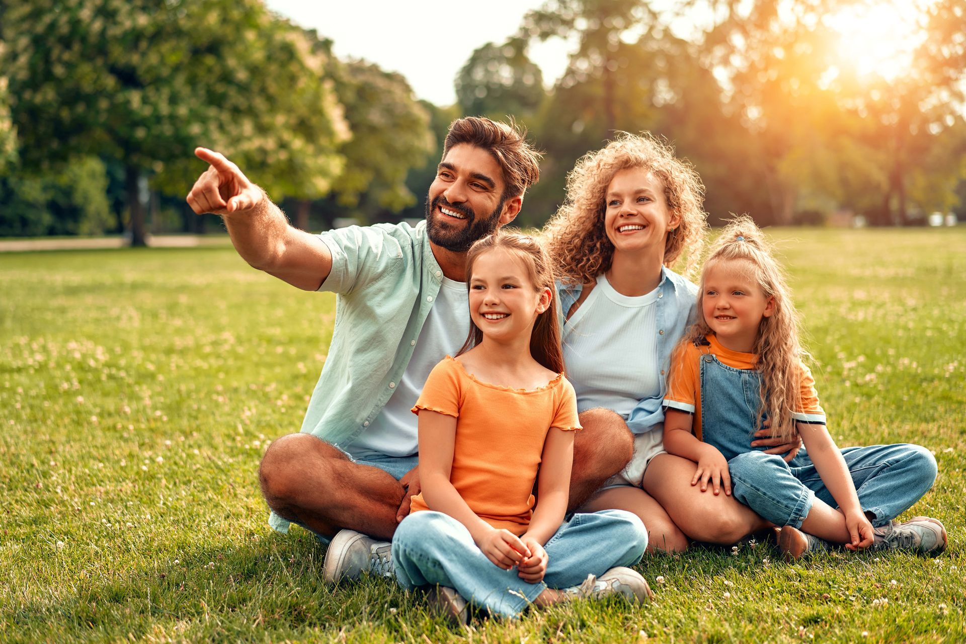 Family of four sitting on grass in a park, smiling. Man points up, sunny background.
