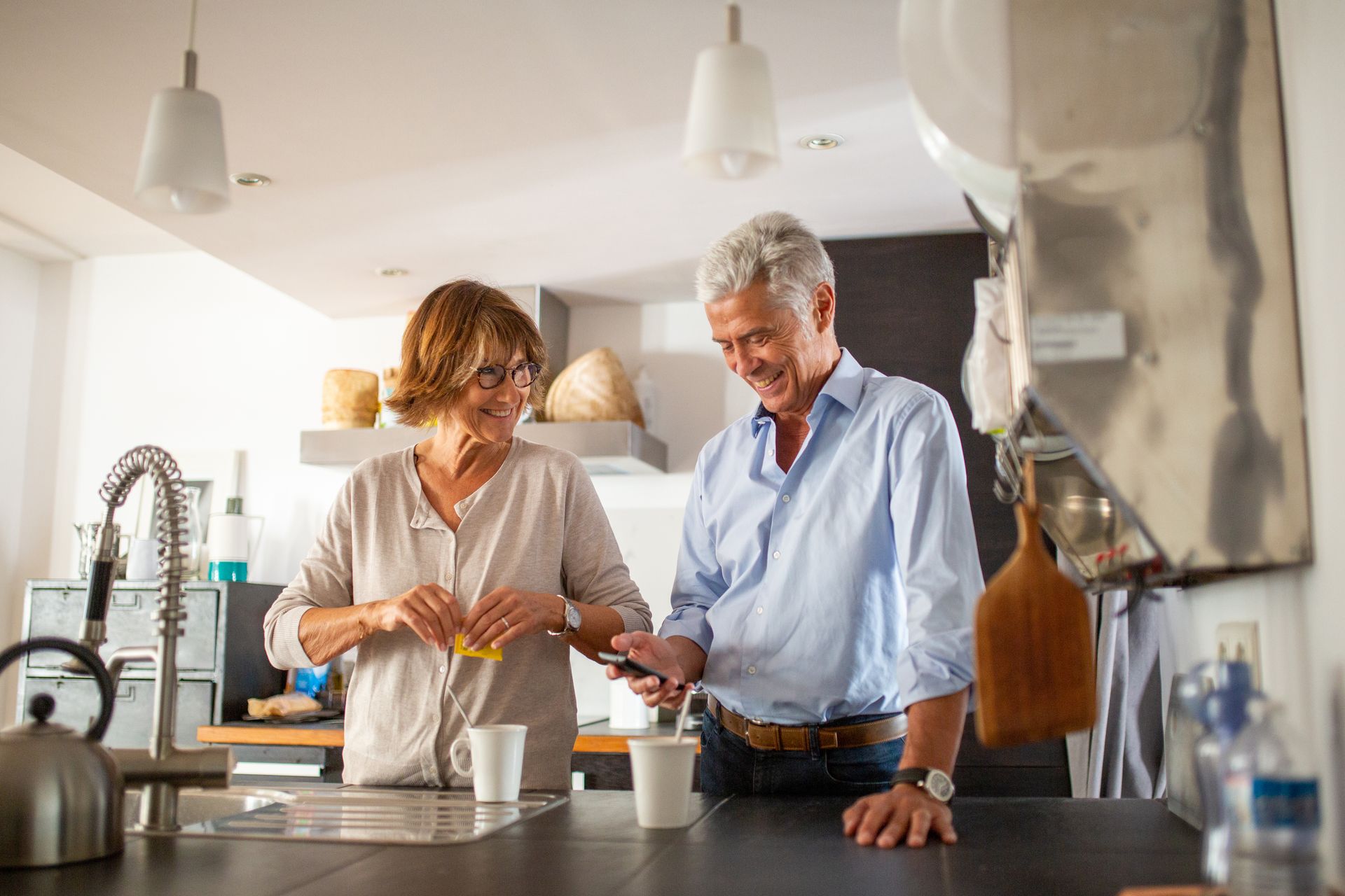 Couple in kitchen, smiling, making coffee. Man holds phone. Woman has glasses, stands by sink.