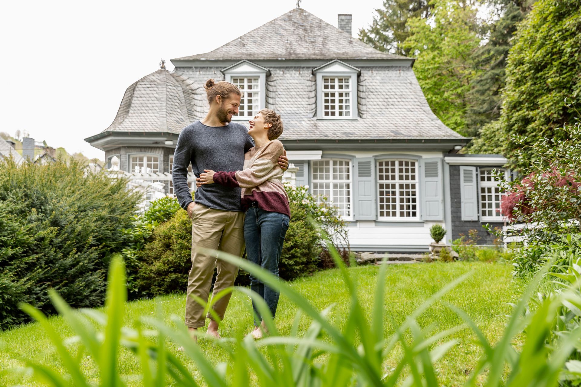 Couple embracing in front of a gray house with a manicured lawn. They are smiling and looking at each other.