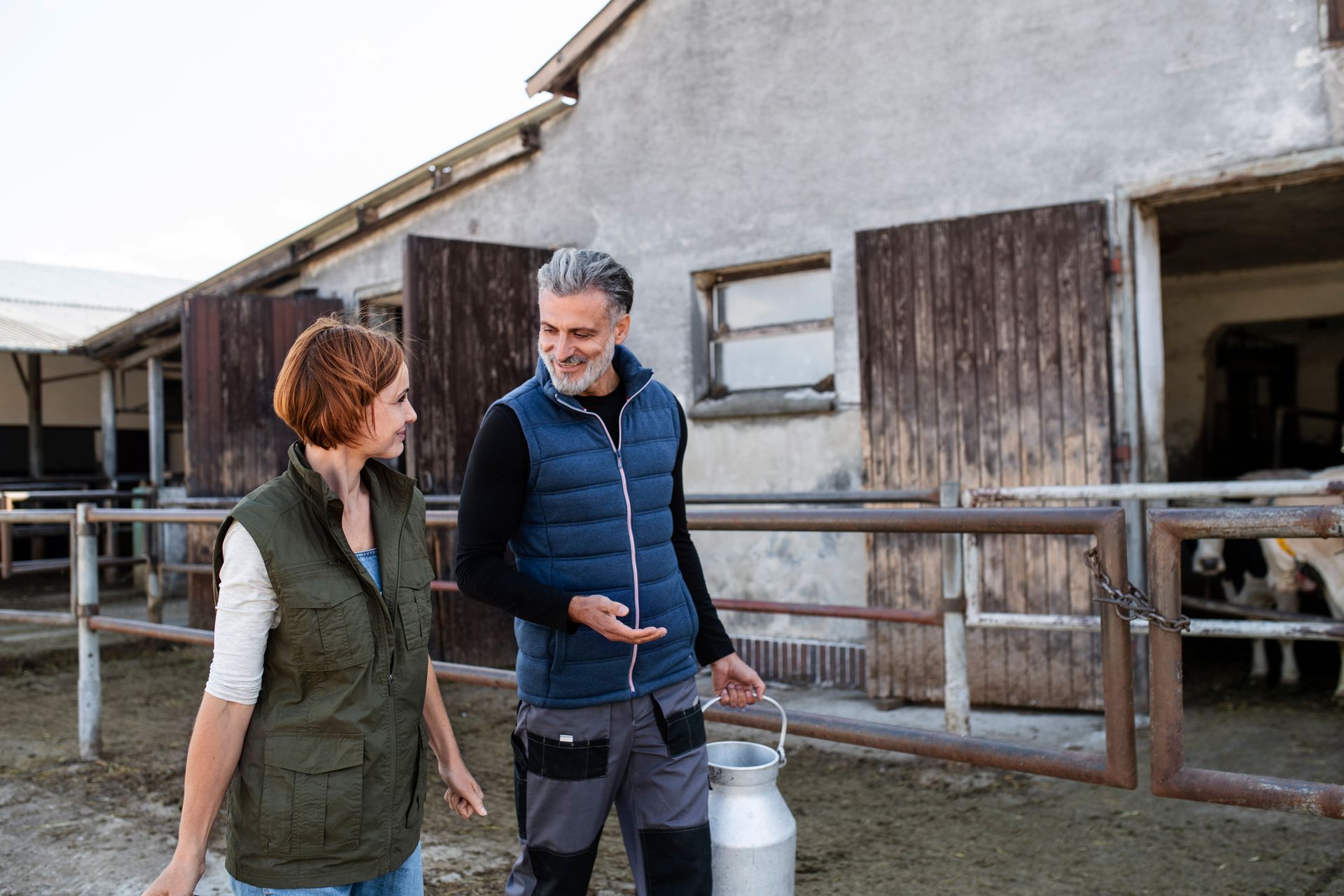 Man and woman in vests walking near a barn, man holding a milk can, smiling.