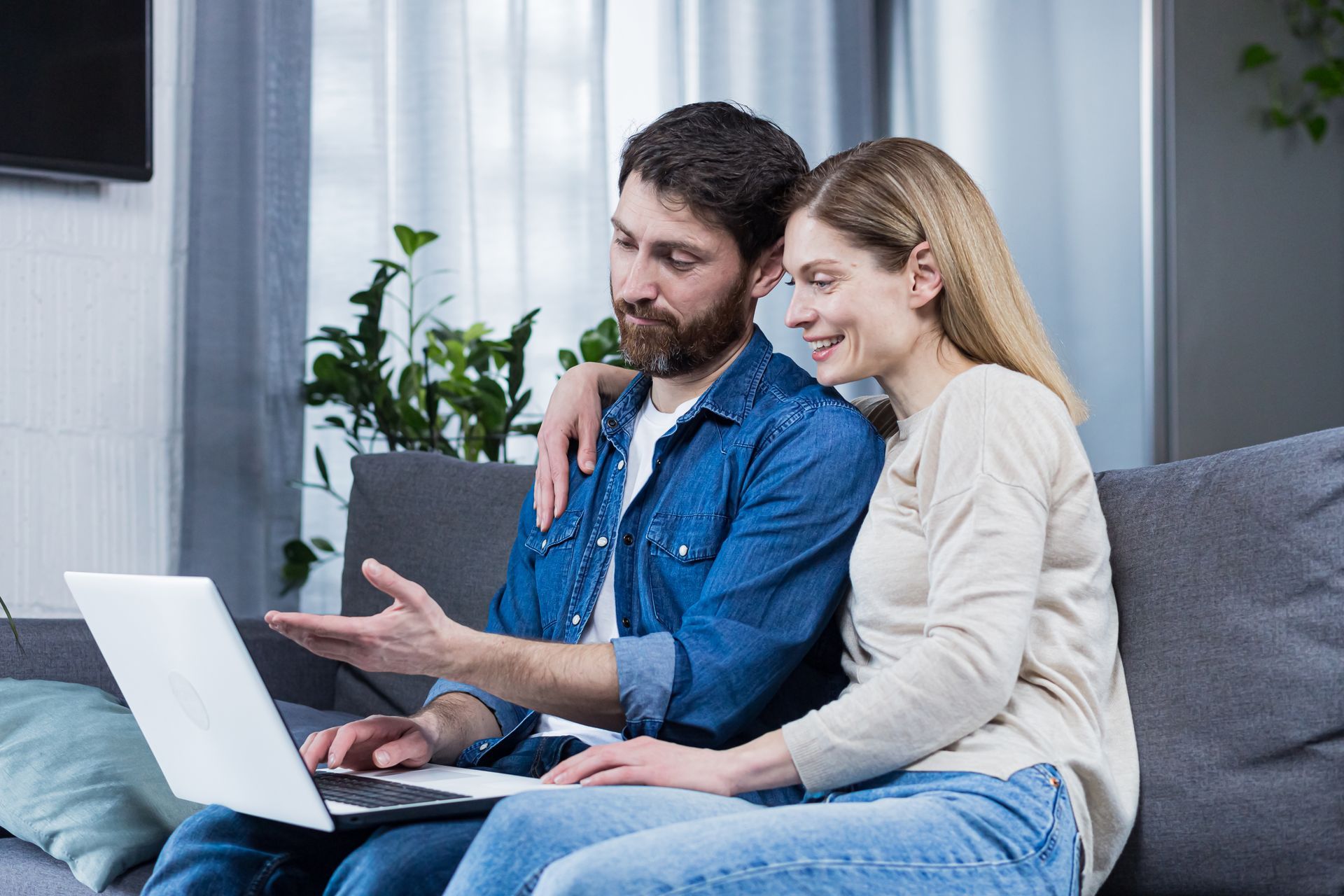 Couple sitting on a couch, looking at a laptop together, man points to screen.